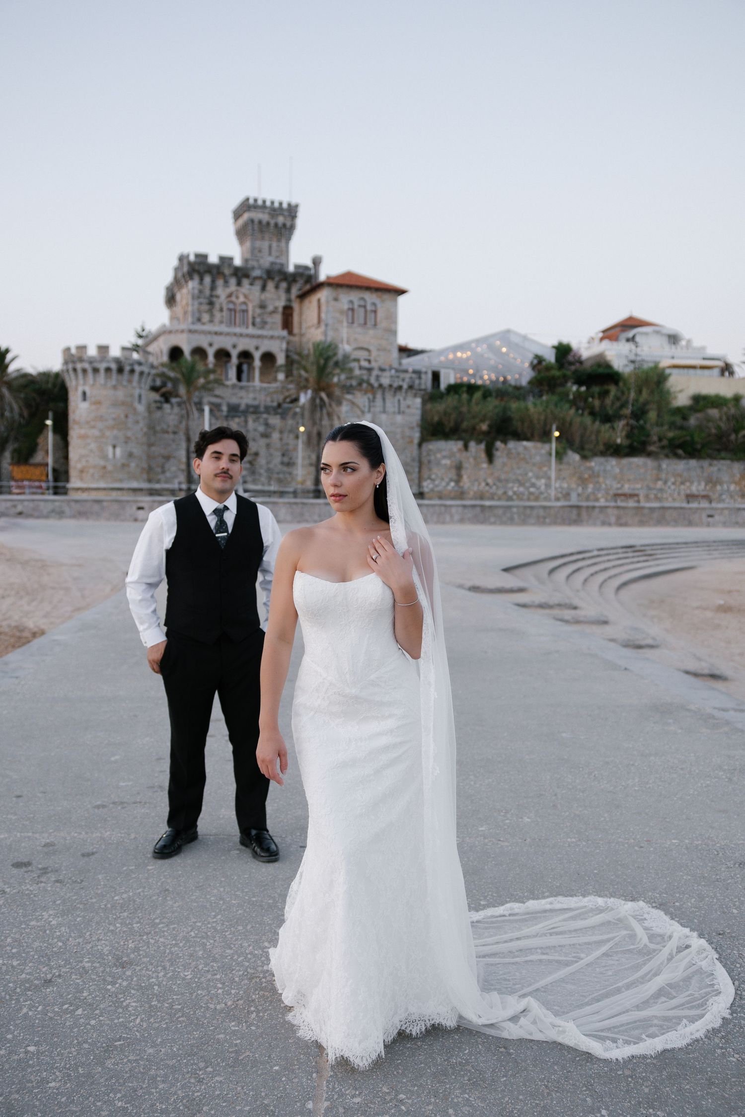 Bride in white gown with veil stands before a castle, groom in black vest behind her at dusk.