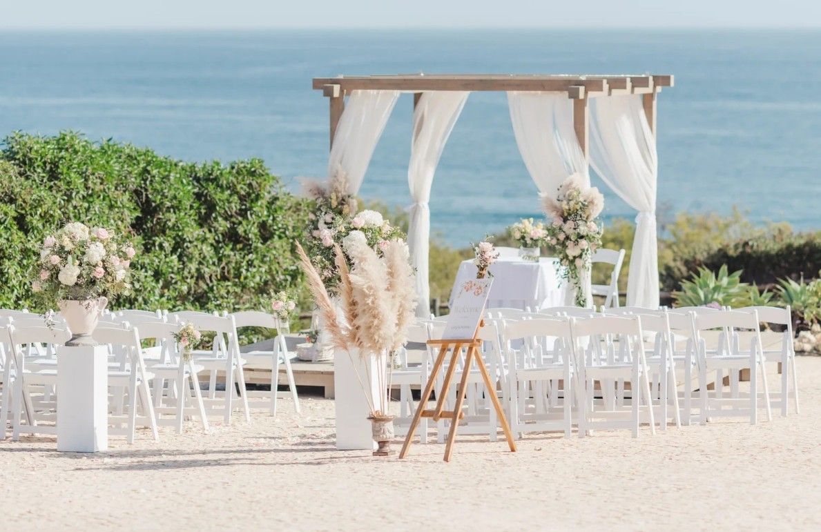Romantic beachside wedding ceremony setup with white chairs, floral arrangements, and draped wooden arch.