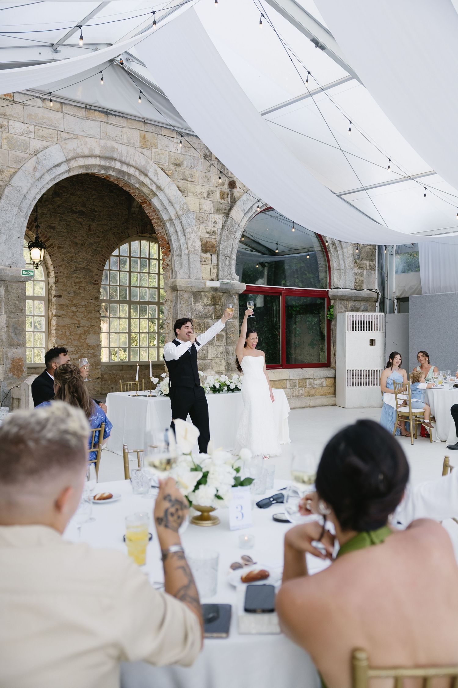 Bride and groom raising glasses during wedding reception toast in elegant stone venue with glass roof.