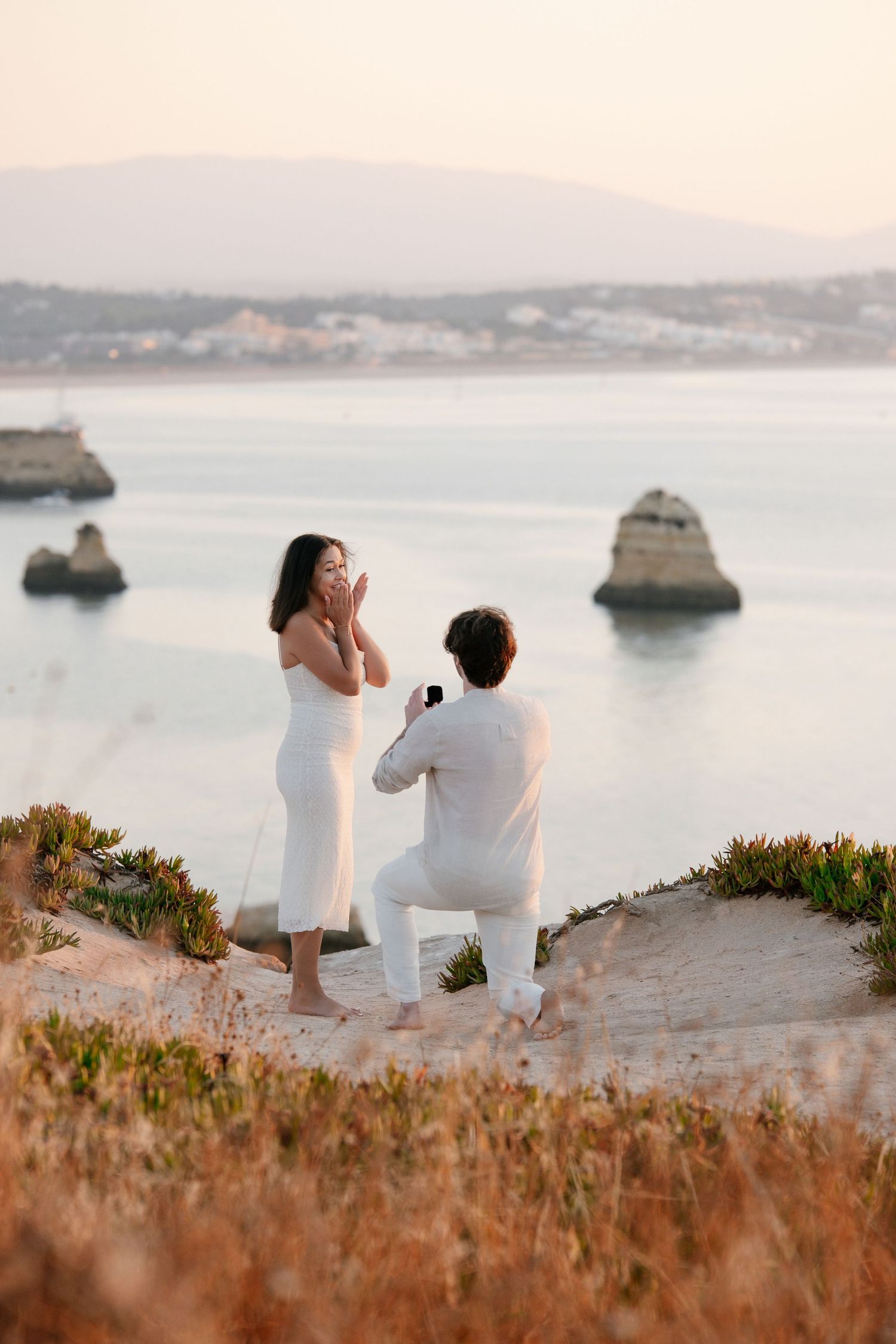 Man proposes on bended knee to surprised woman in white dress on scenic coastal cliffside at golden hour.