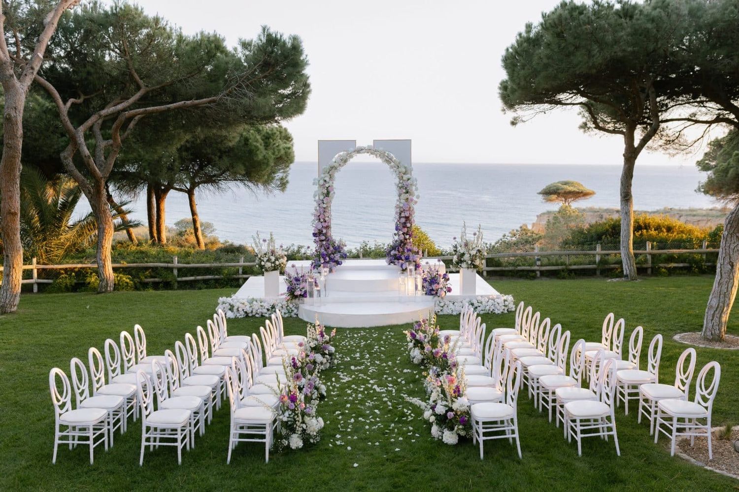 Beautiful outdoor wedding ceremony setup with floral arch, white chairs, and ocean views at sunset.