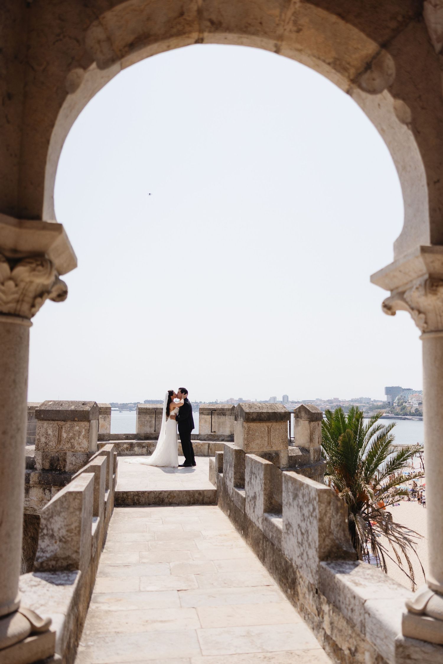 Bride and groom share a romantic kiss framed by a stone arch on a castle terrace overlooking the sea.
