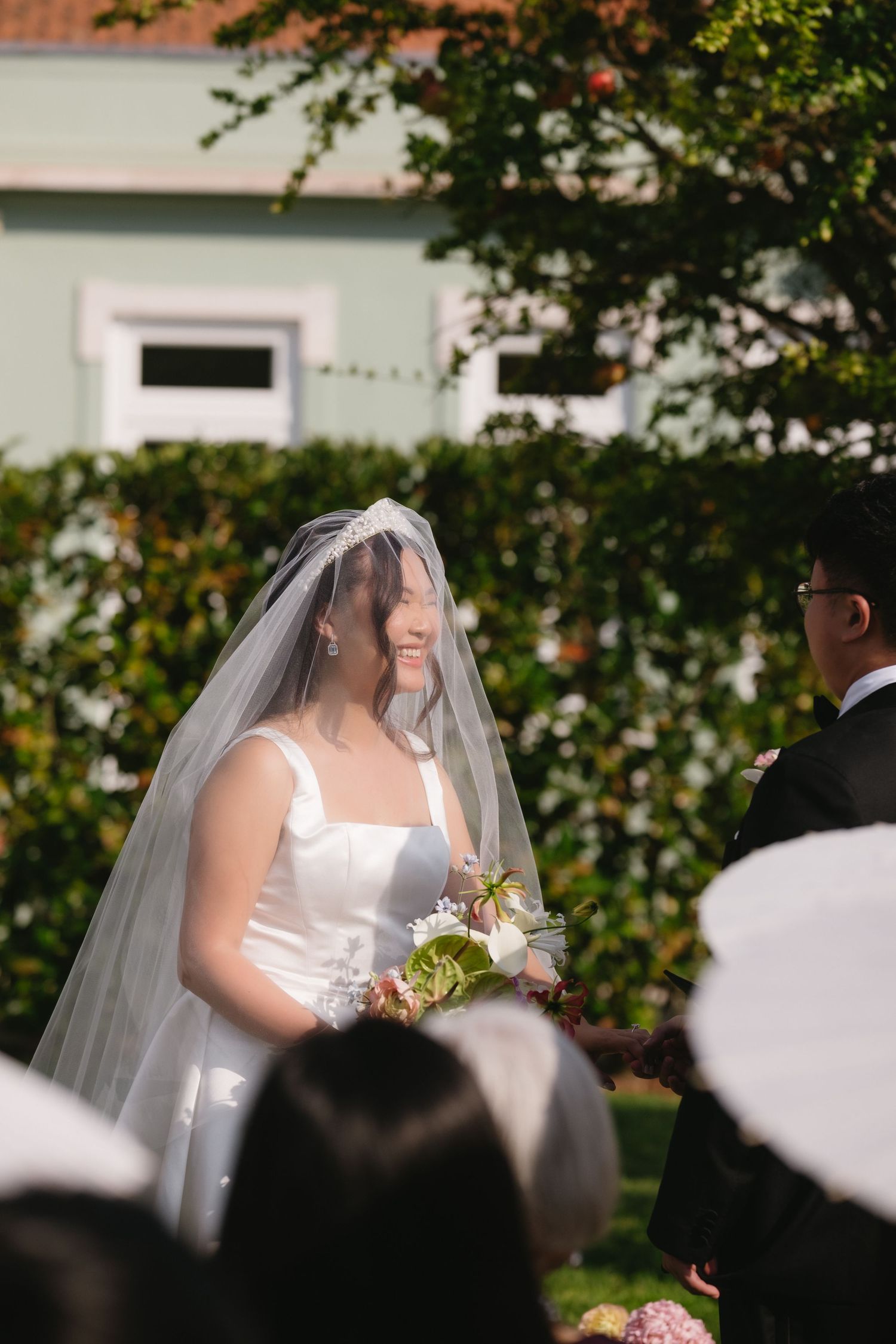 Smiling Asian bride in white gown and veil holding bouquet at outdoor wedding ceremony with lush green hedges.