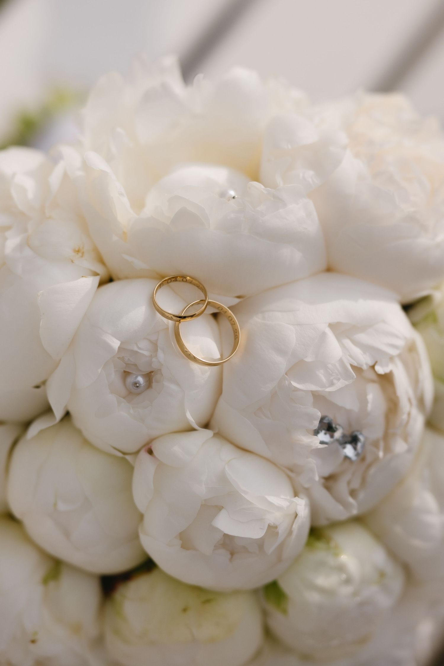 Two gold wedding rings resting on a bridal bouquet of white peonies with pearl and crystal accents.