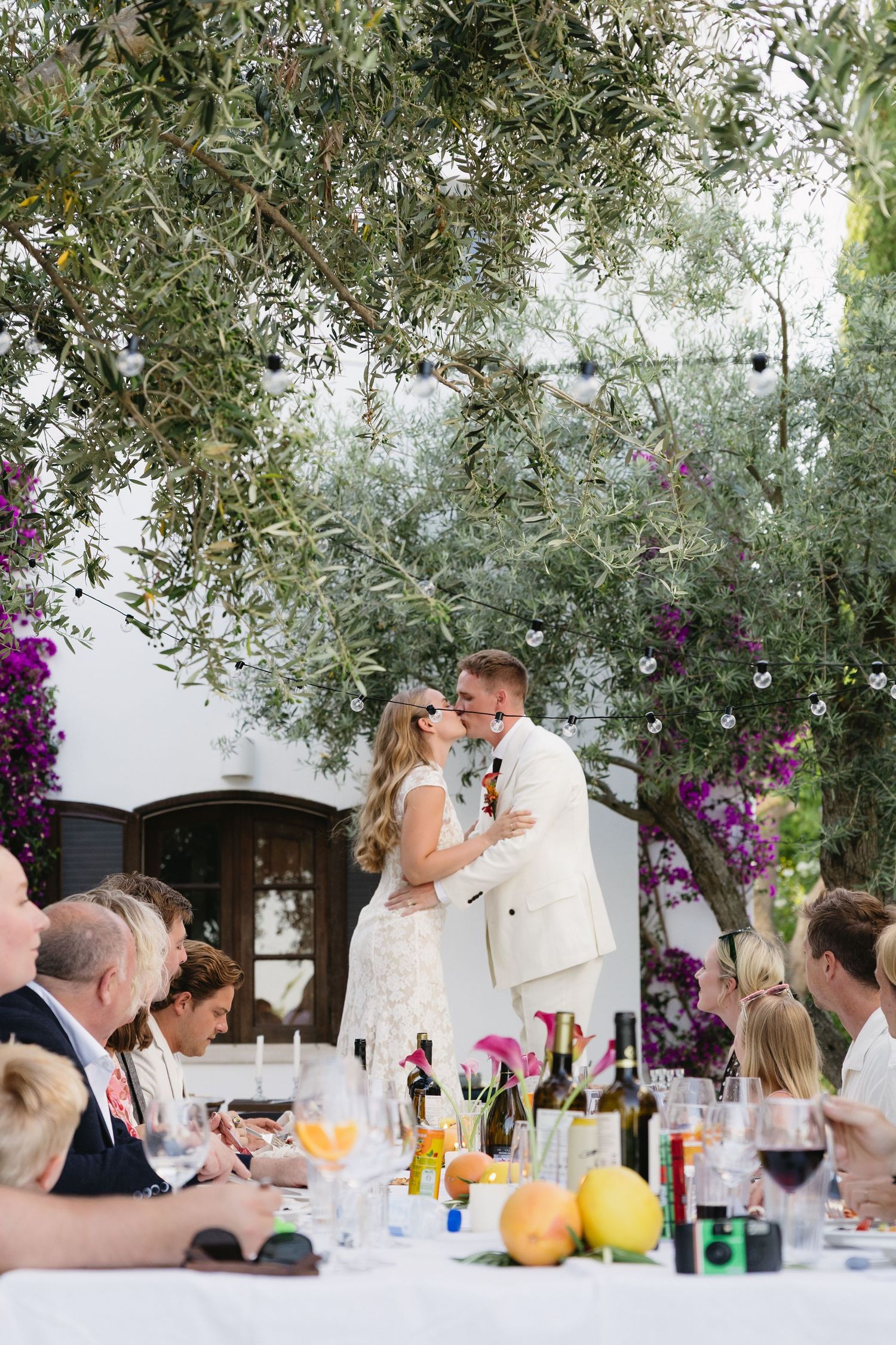 Newlyweds share a kiss at outdoor reception table under olive tree with string lights and bougainvillea.