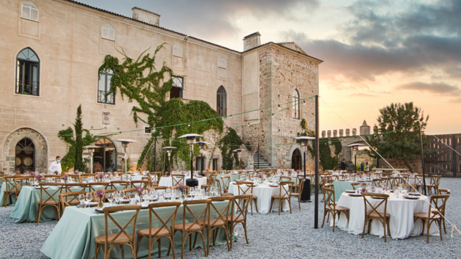 Elegant outdoor wedding reception setup in a historic stone castle courtyard with long tables and wooden chairs at sunset.