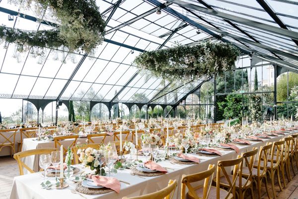 Elegant greenhouse wedding reception with long tables, floral arrangements, and natural light through glass ceiling.