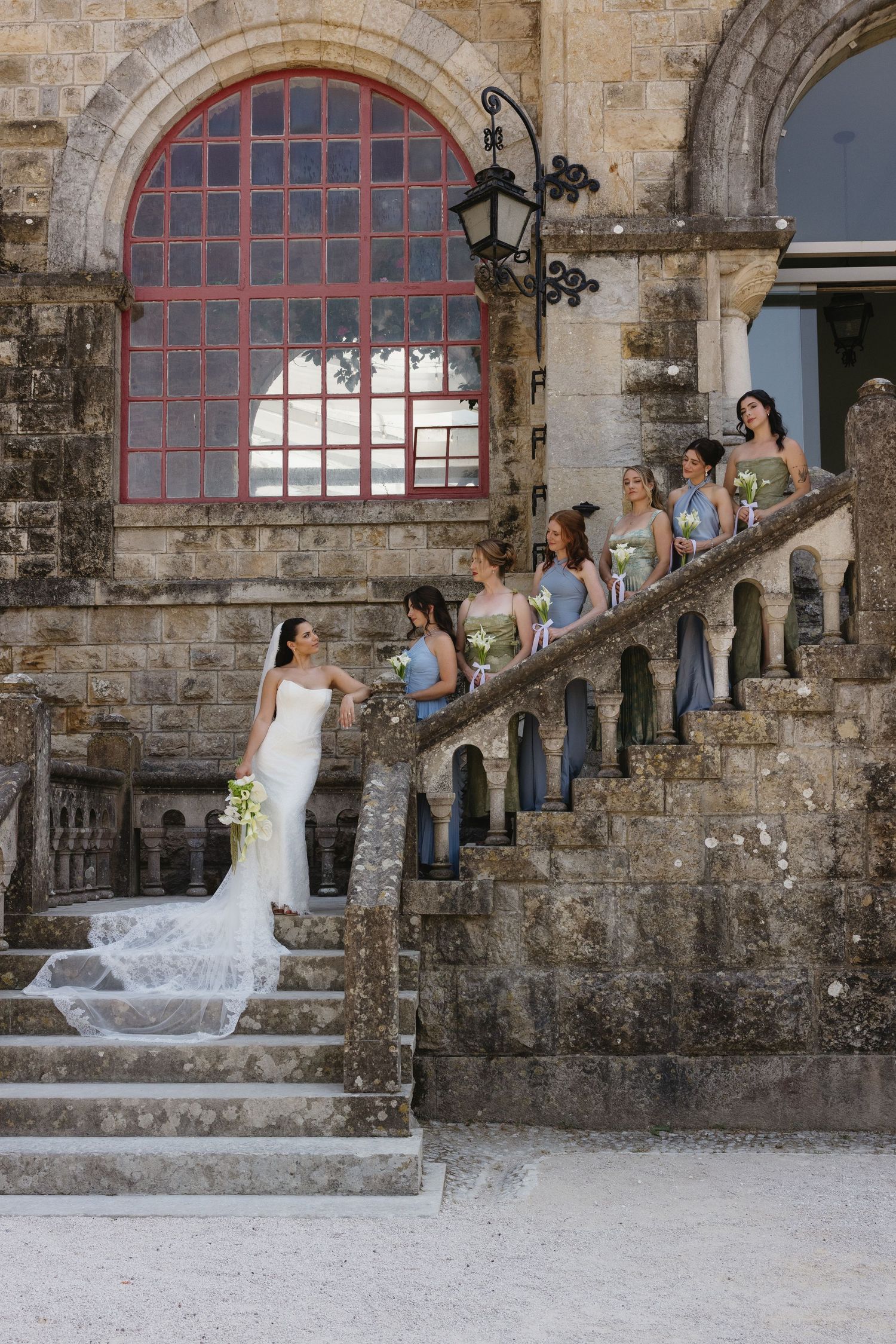 Bride in white gown with bridesmaids in gray dresses posing on stone staircase of historic venue.