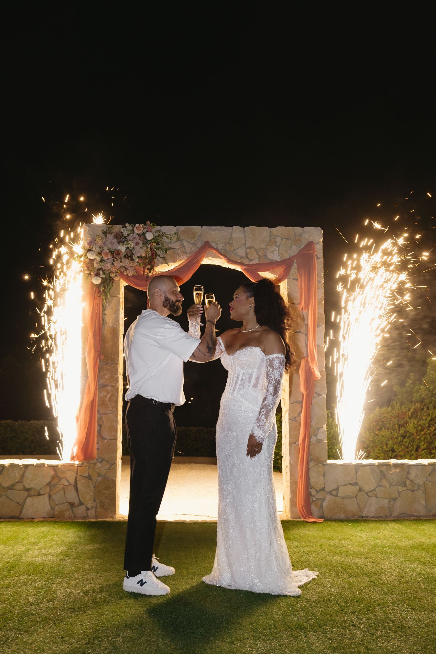 Newlyweds toasting champagne at night with dramatic sparkler fountains behind floral arch at wedding reception.