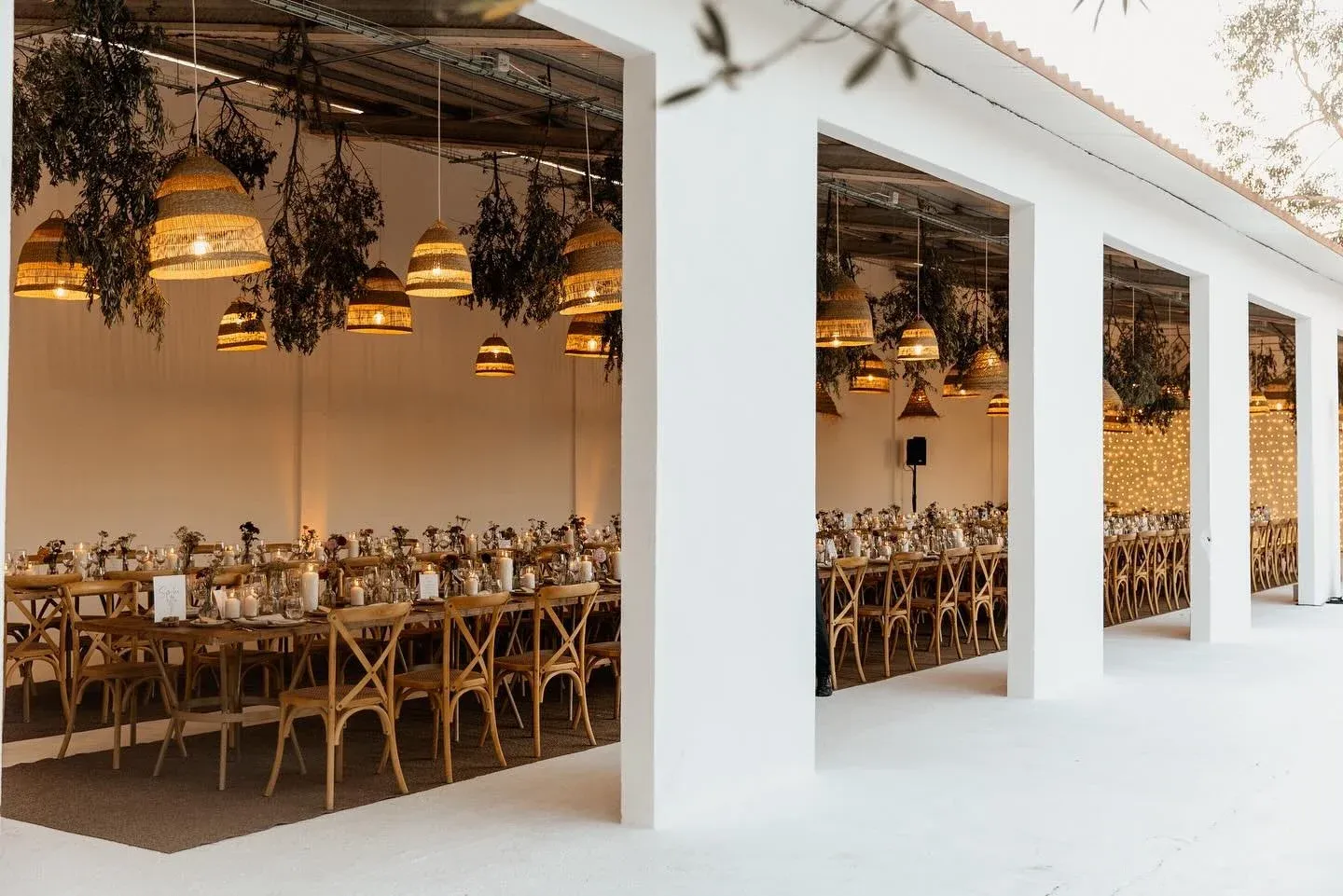 Elegant wedding reception venue with long wooden tables, rattan pendant lights, and hanging dried botanicals.