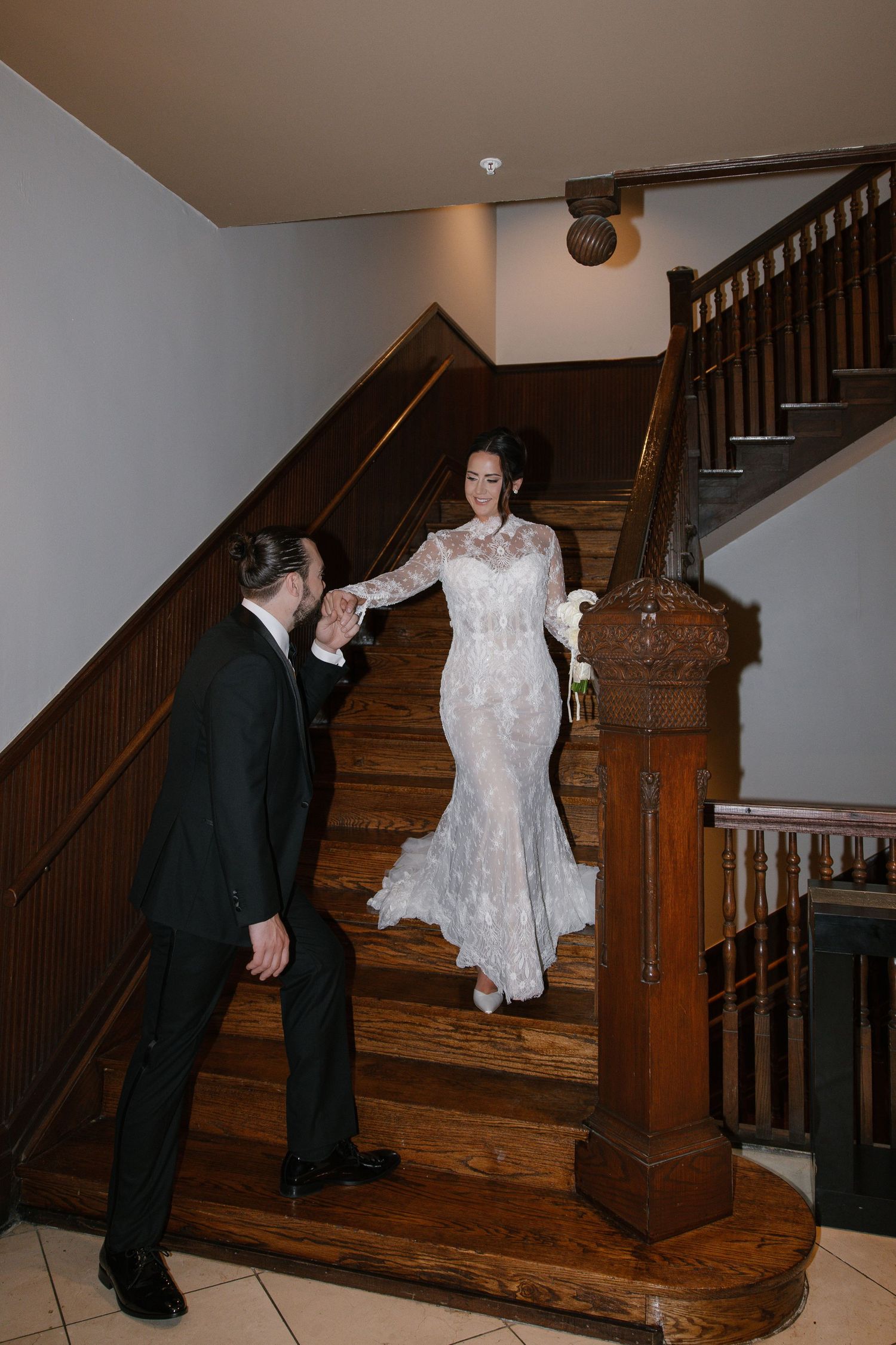 Bride in lace gown descends wooden staircase as groom reaches for her hand during wedding day portraits.