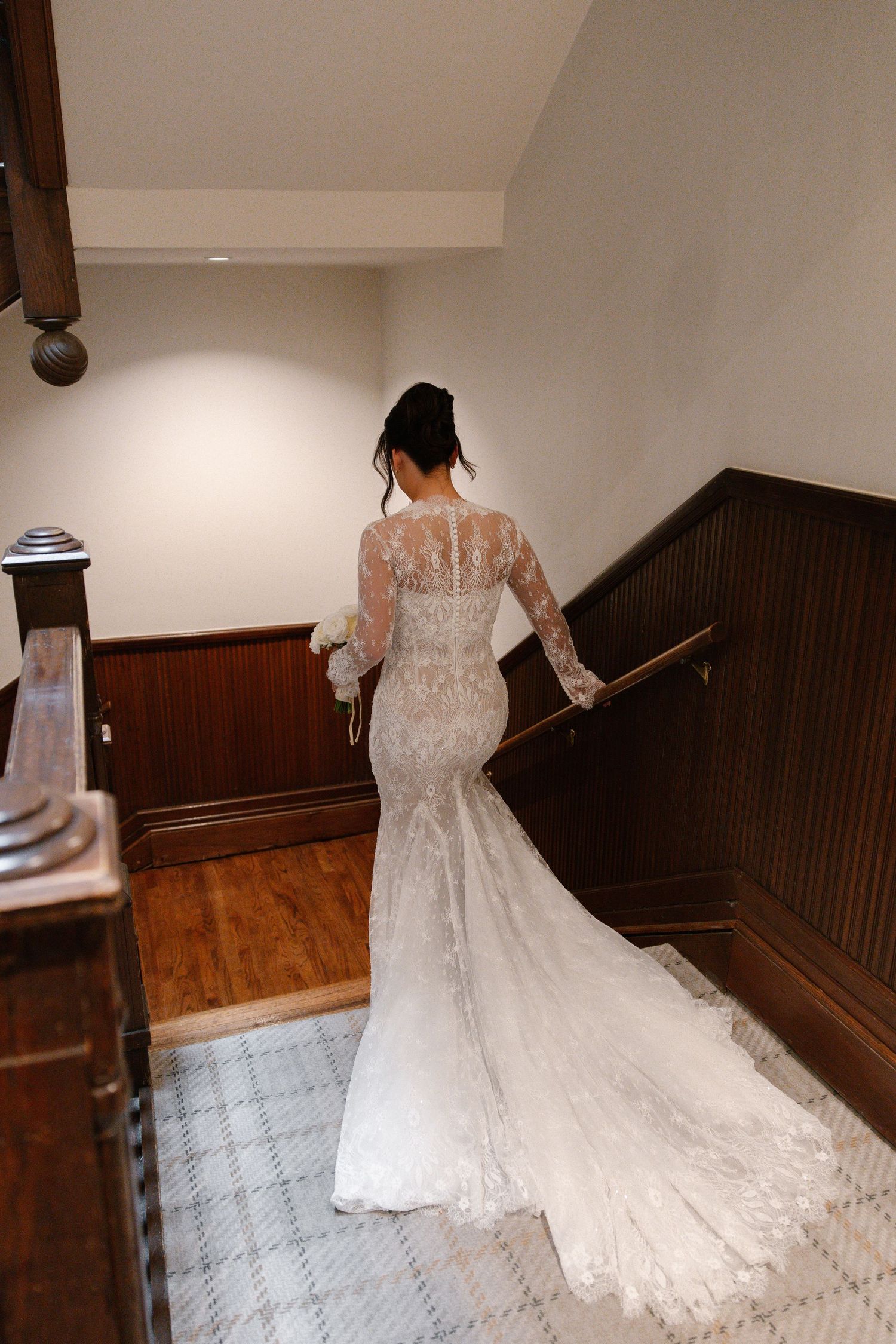 Bride in lace long-sleeve mermaid wedding gown with train walks up wooden staircase, viewed from behind.