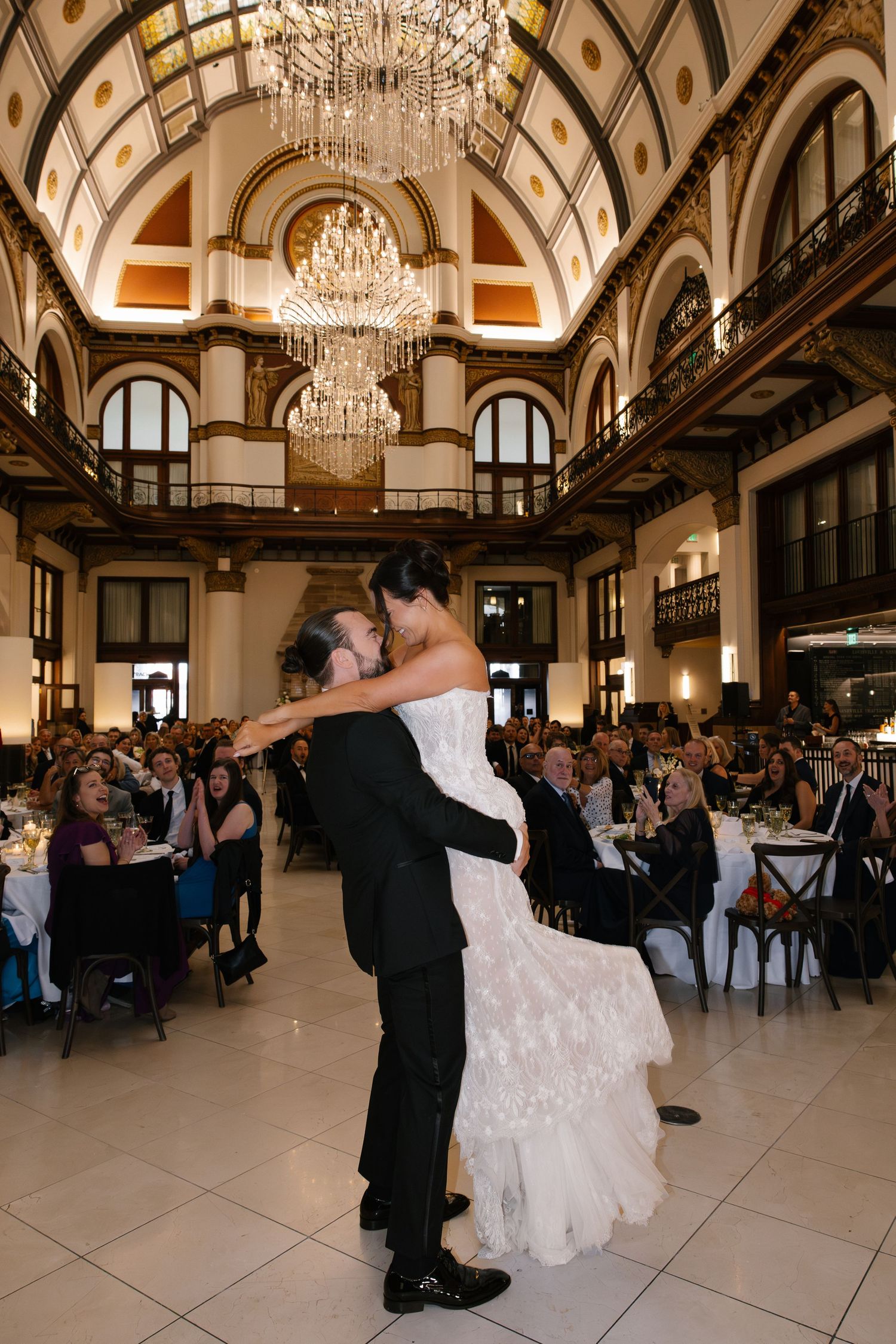 Groom lifts bride during first dance in elegant ballroom with crystal chandeliers and guests seated at reception tables.