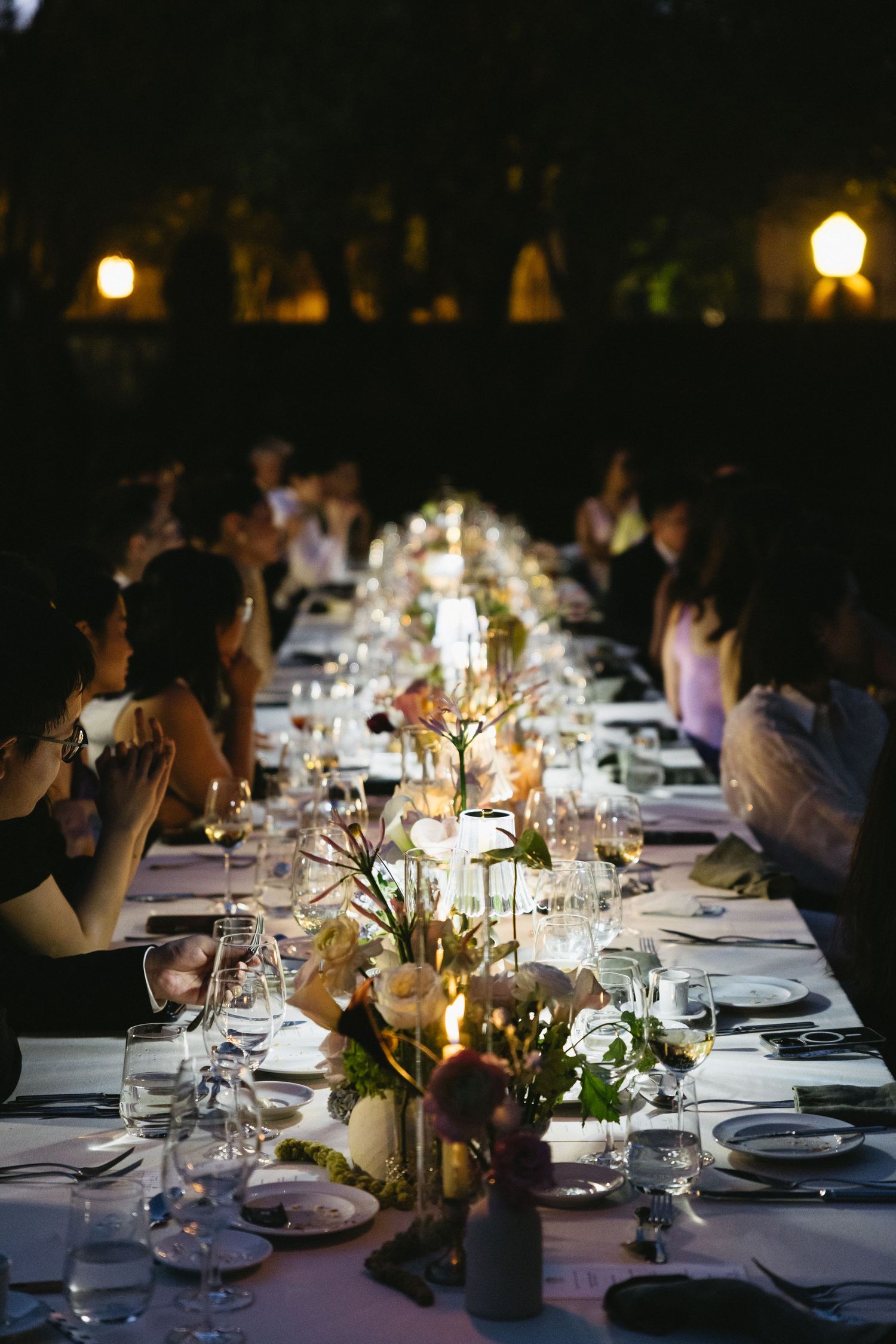 Elegant candlelit outdoor dinner party with long banquet table, floral centerpieces, and guests dining under evening sky.