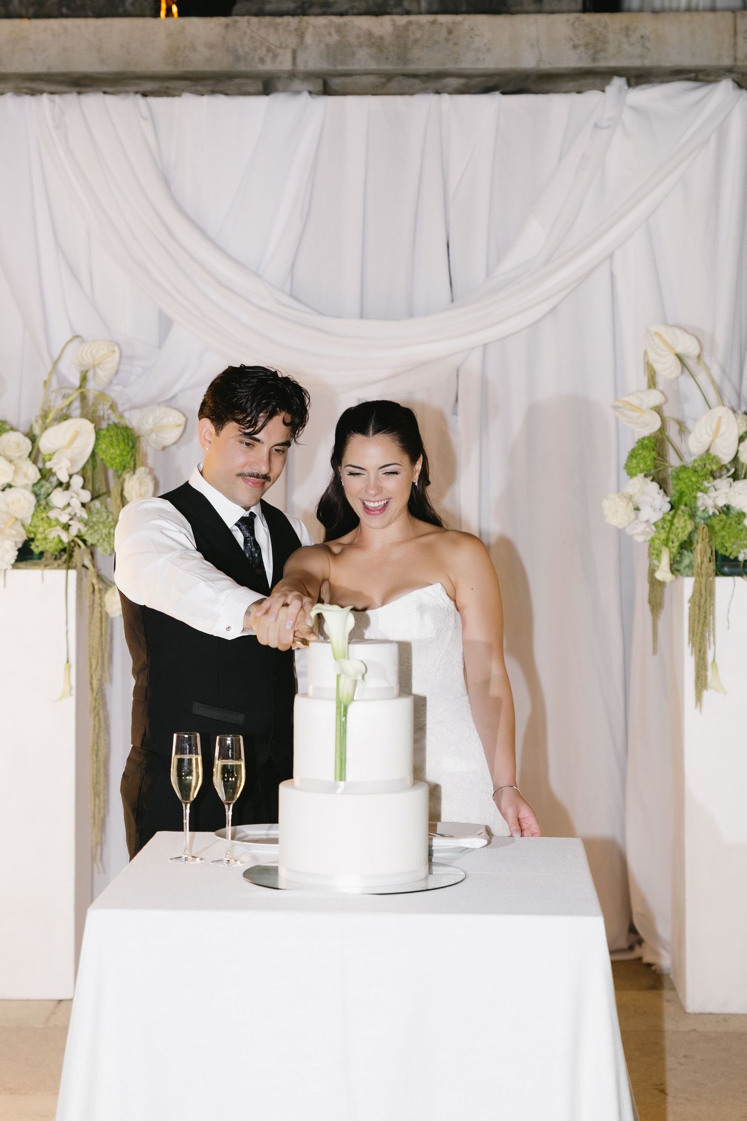 Bride and groom cutting white three-tier wedding cake at elegant reception with champagne flutes and floral arrangements.