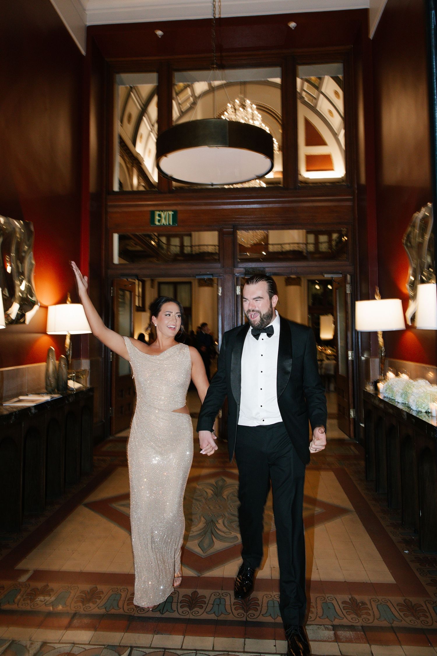 Elegant couple in formal attire walking hand-in-hand through a grand hotel lobby, woman waving in a sequined gown.