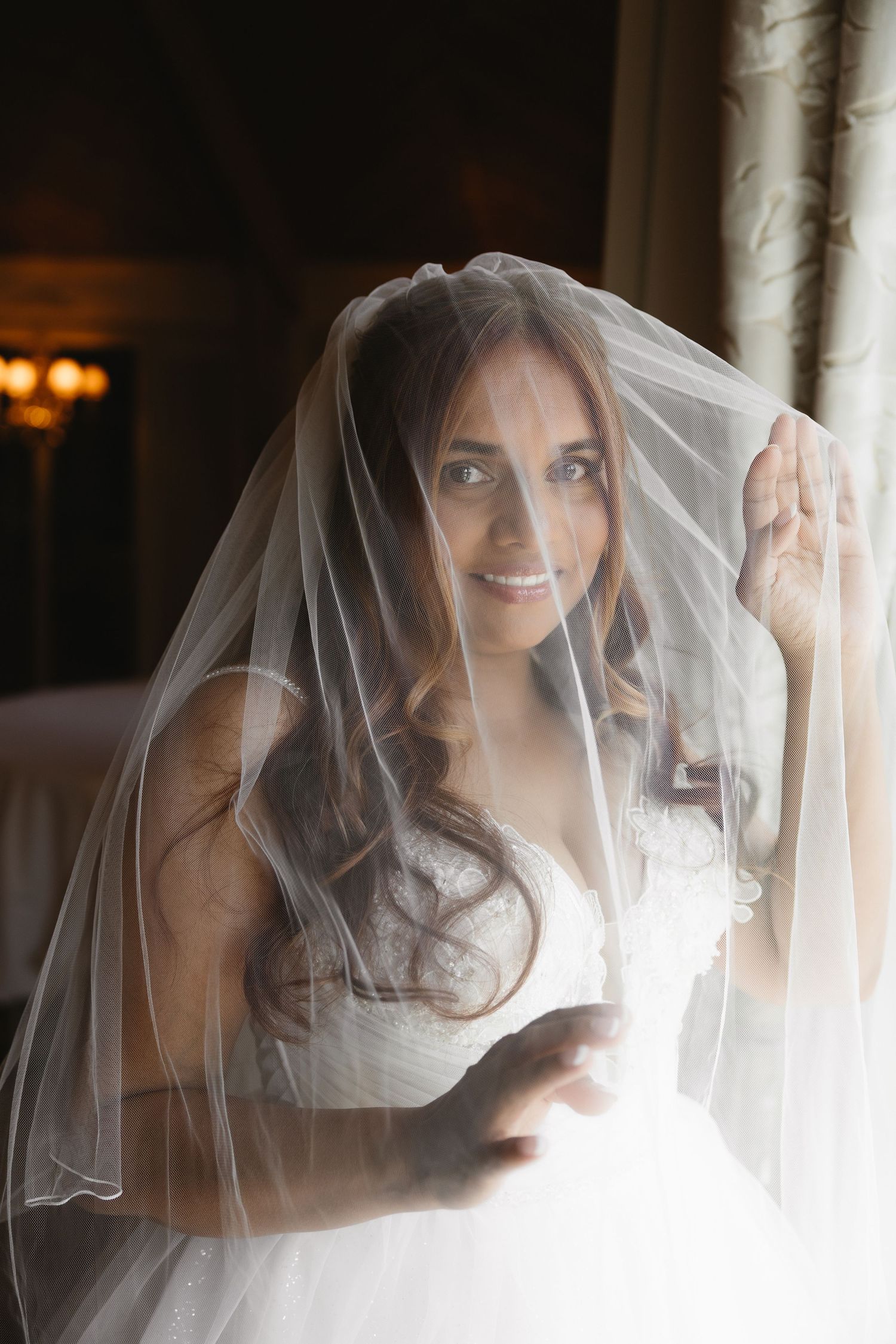 Smiling bride in white wedding dress and veil lifted over her face, posing near a window in elegant indoor setting.