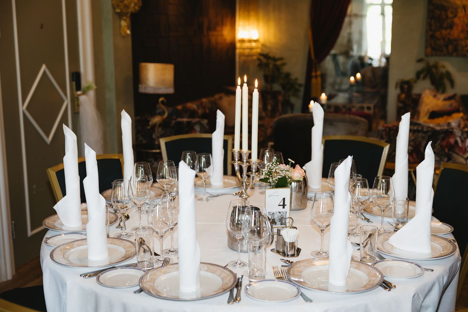 Elegant round dining table set with white taper candles, crystal glasses, and gold-rimmed plates for a formal event.