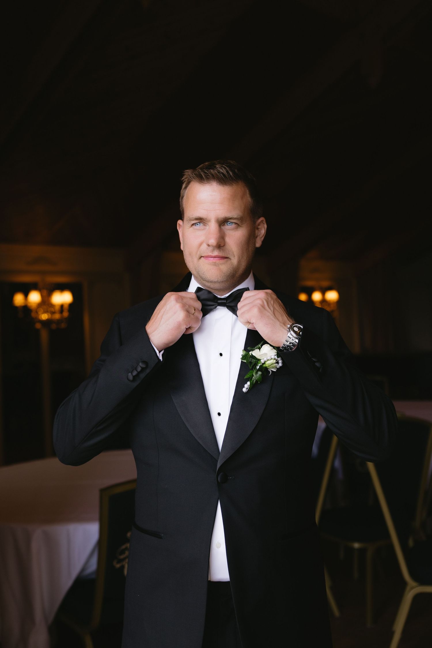 Groom in black tuxedo adjusting bow tie with boutonniere, posing in elegant dimly lit wedding venue.