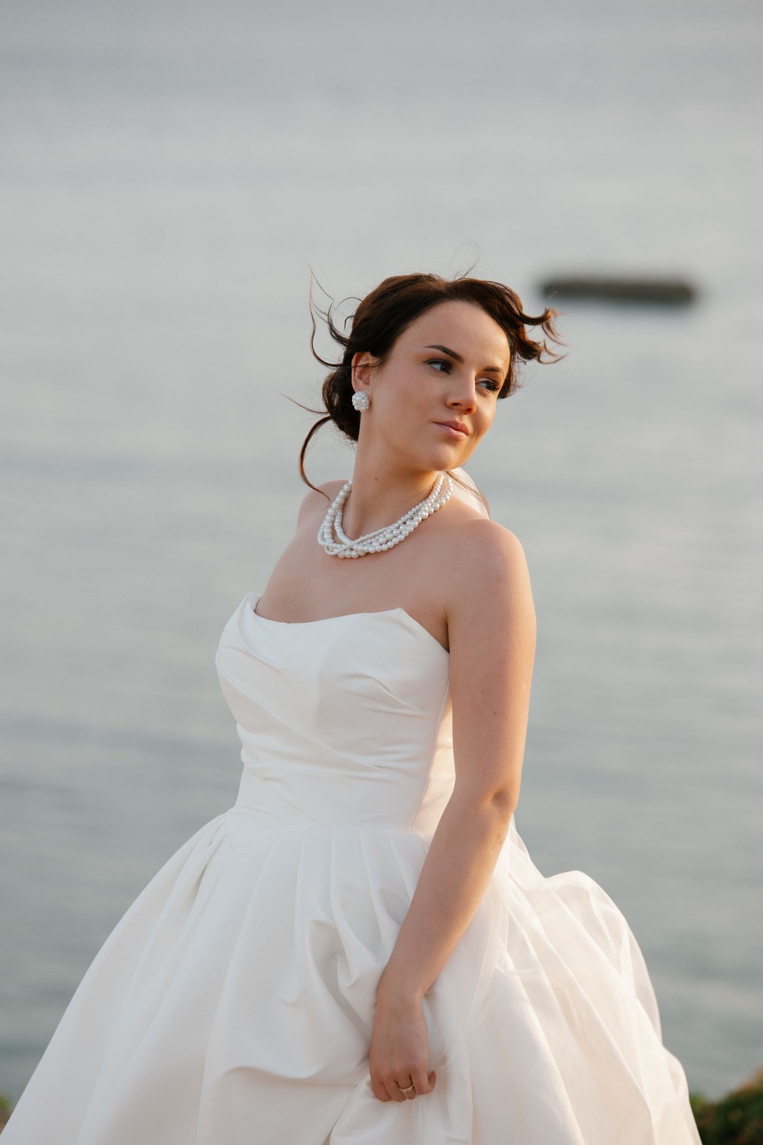 Beautiful bride in strapless white gown and pearl necklace stands by the ocean with wind in her hair.