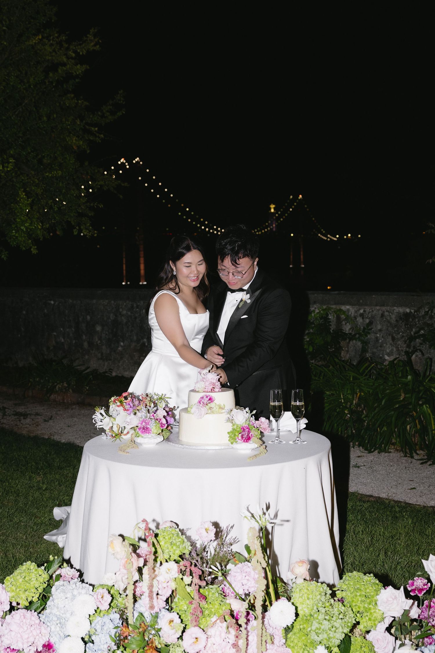 Bride and groom cutting their floral-adorned wedding cake outdoors at night under string lights.