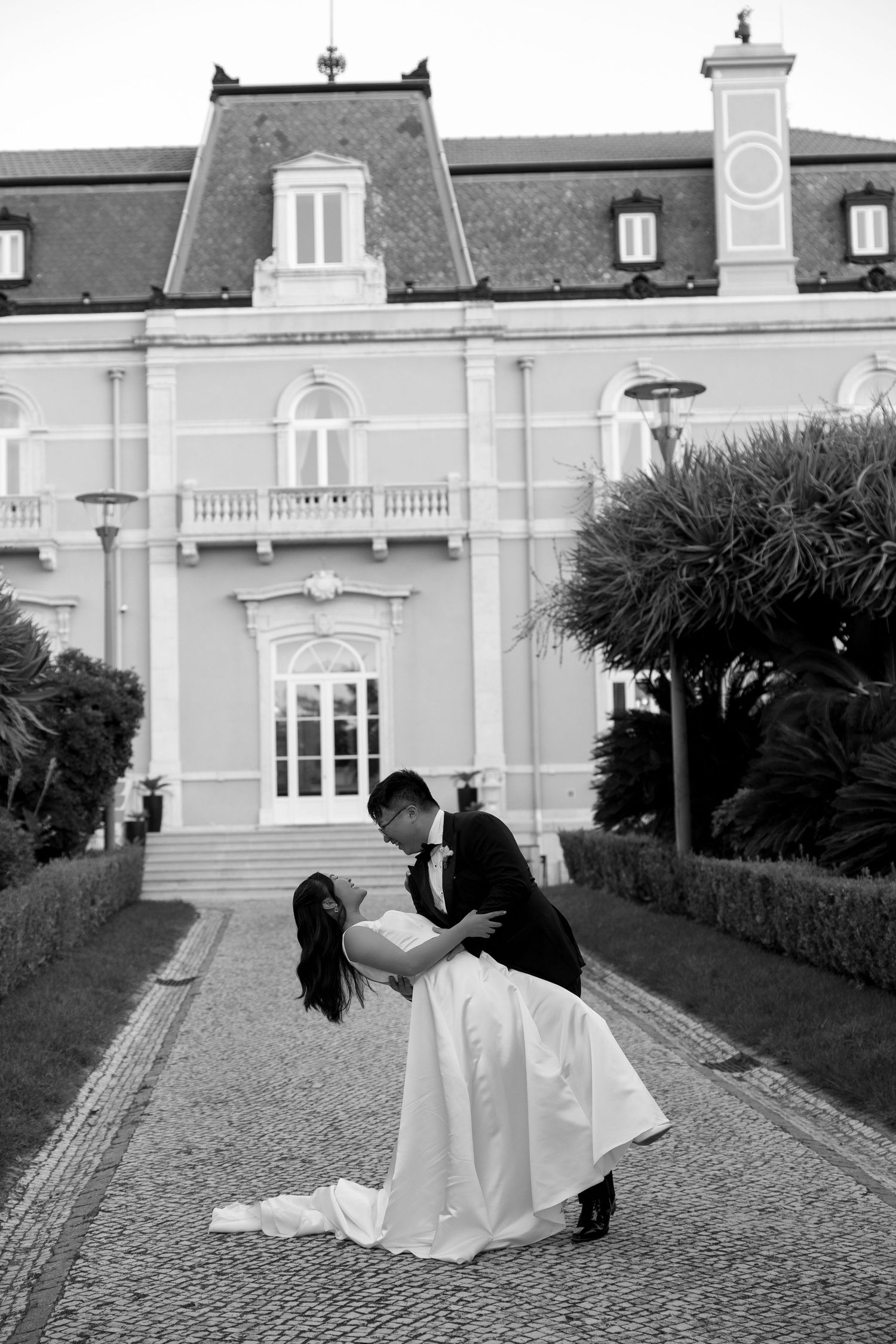 Black and white photo of a groom dipping his bride in front of an elegant mansion on their wedding day.