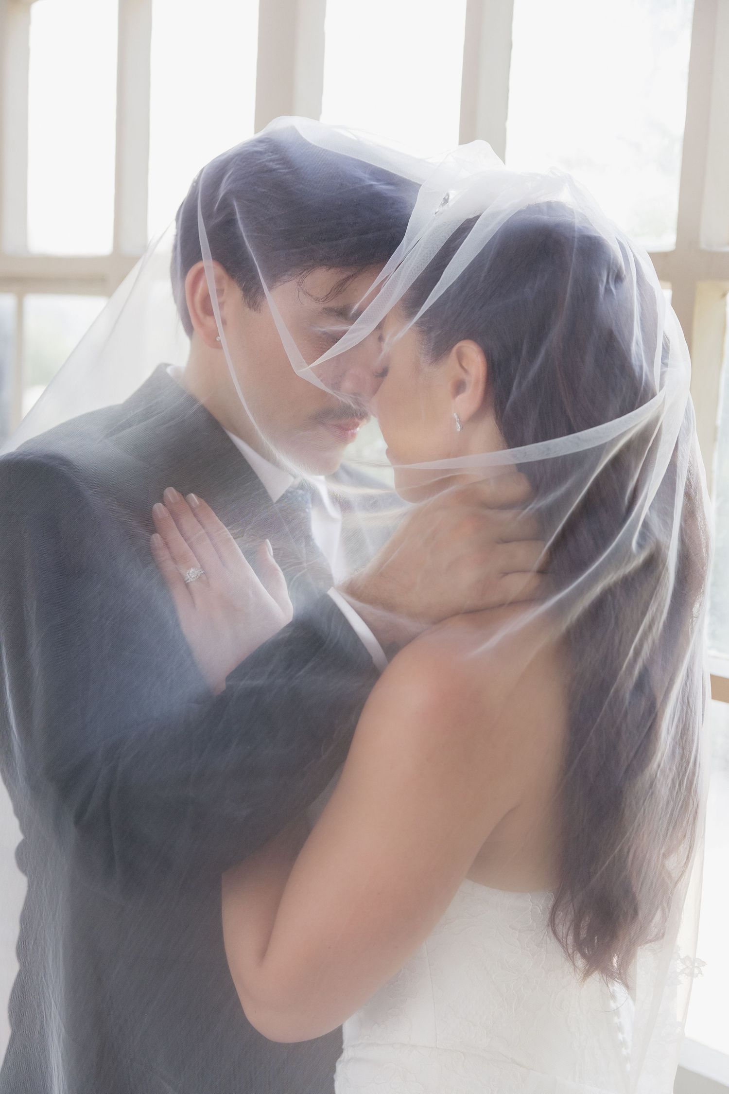 Bride and groom share a romantic kiss under a flowing white veil on their wedding day.