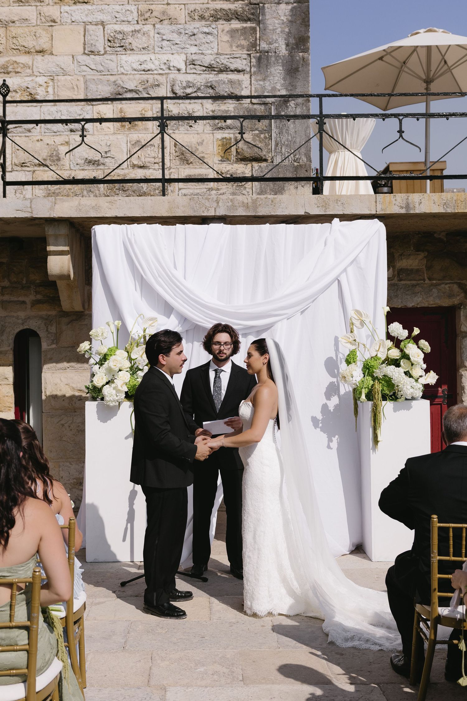 Bride and groom exchange vows at outdoor wedding ceremony beneath white draped arch with floral arrangements.