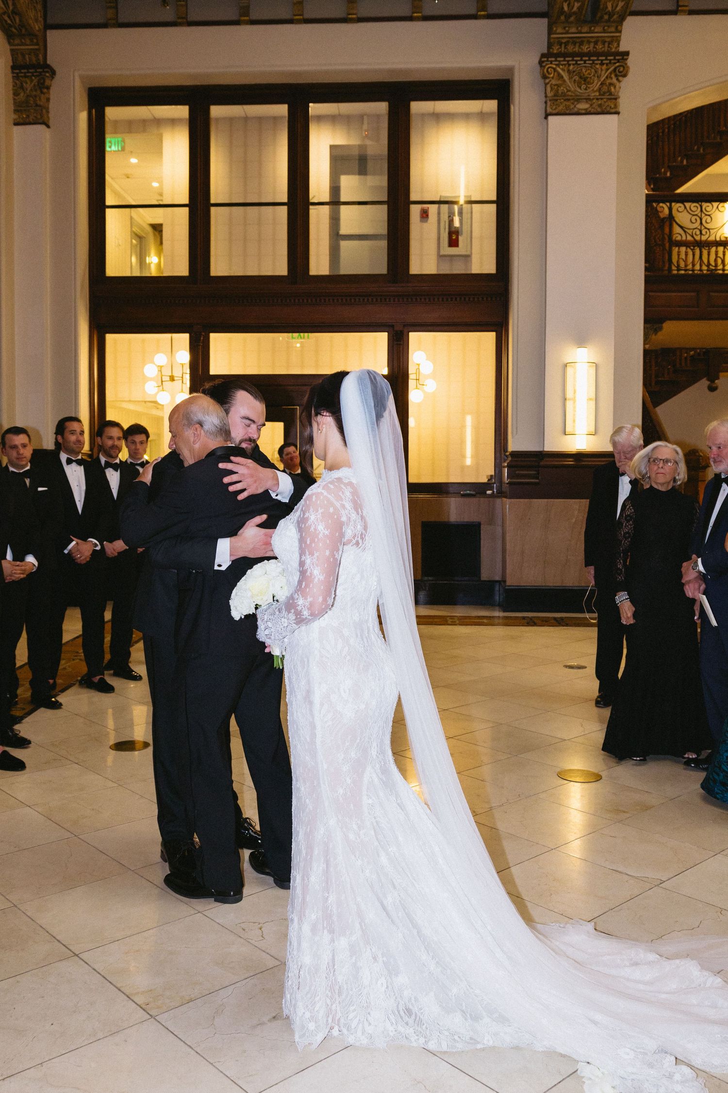 Bride in lace gown and veil embraces family during elegant wedding ceremony in grand ballroom venue.