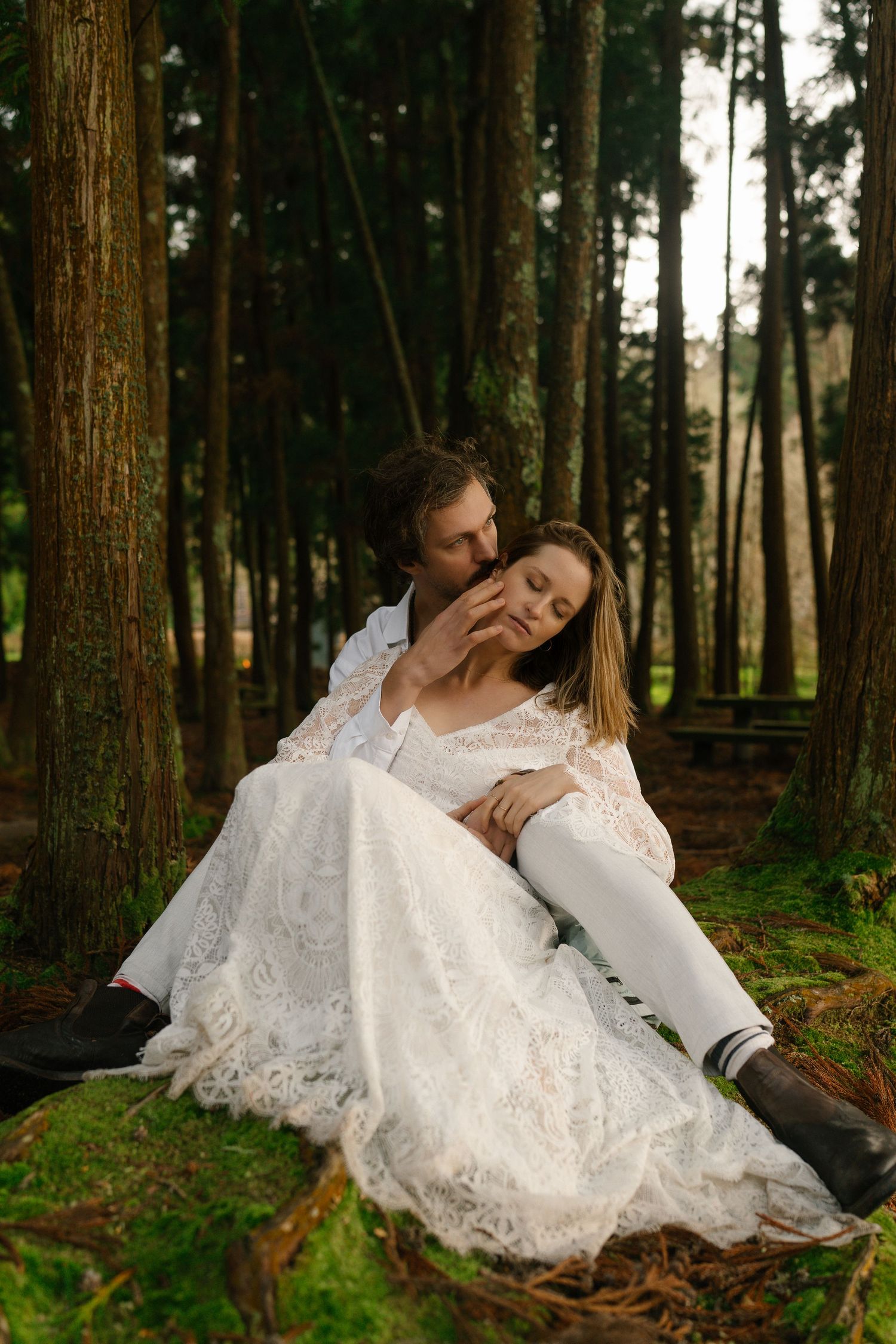 Groom tenderly kissing bride as she reclines in his arms amid tall trees in a lush forest setting.