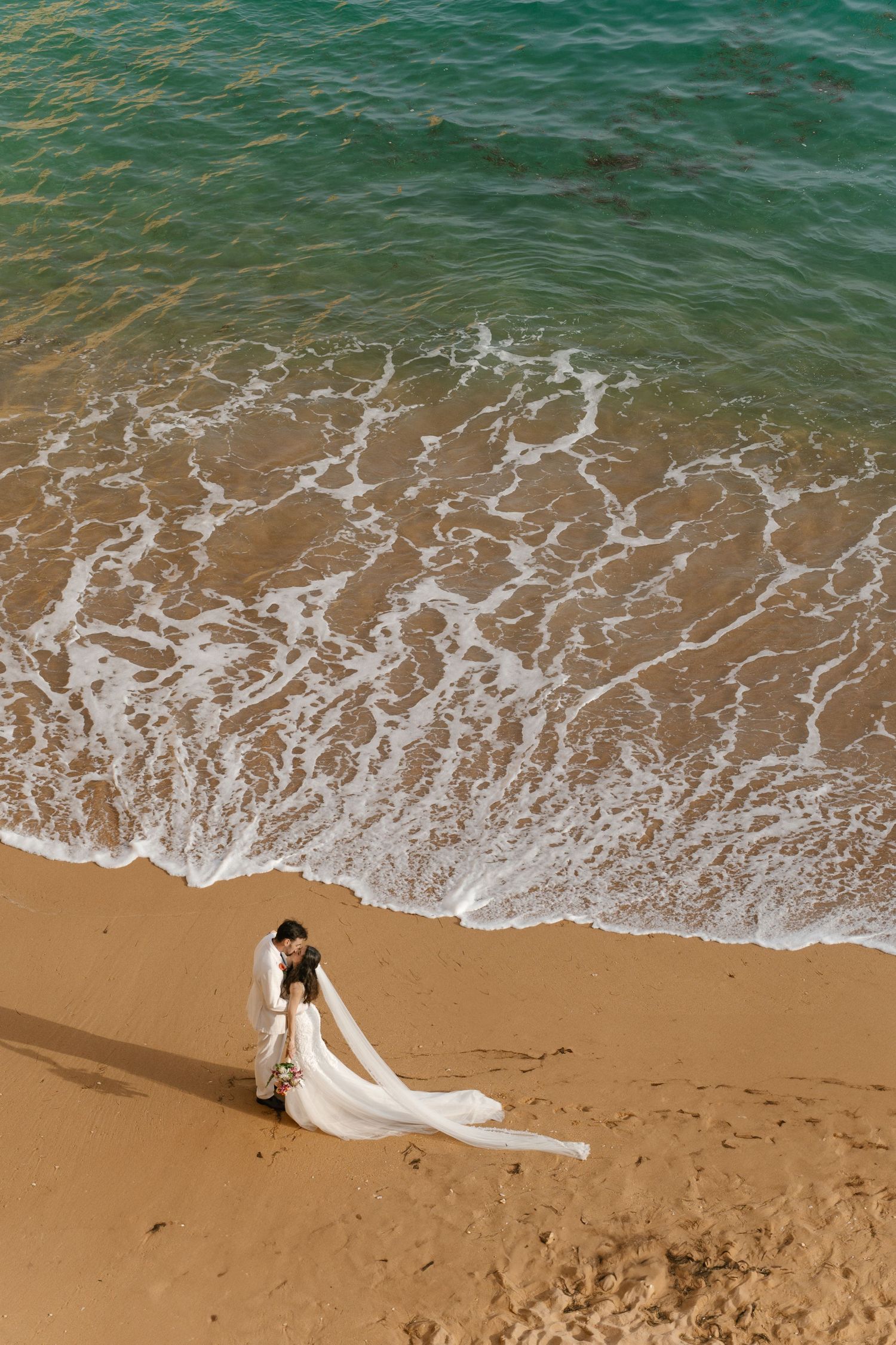 Bride and groom sharing a tender moment on the sandy beach with ocean waves creating a dreamy backdrop