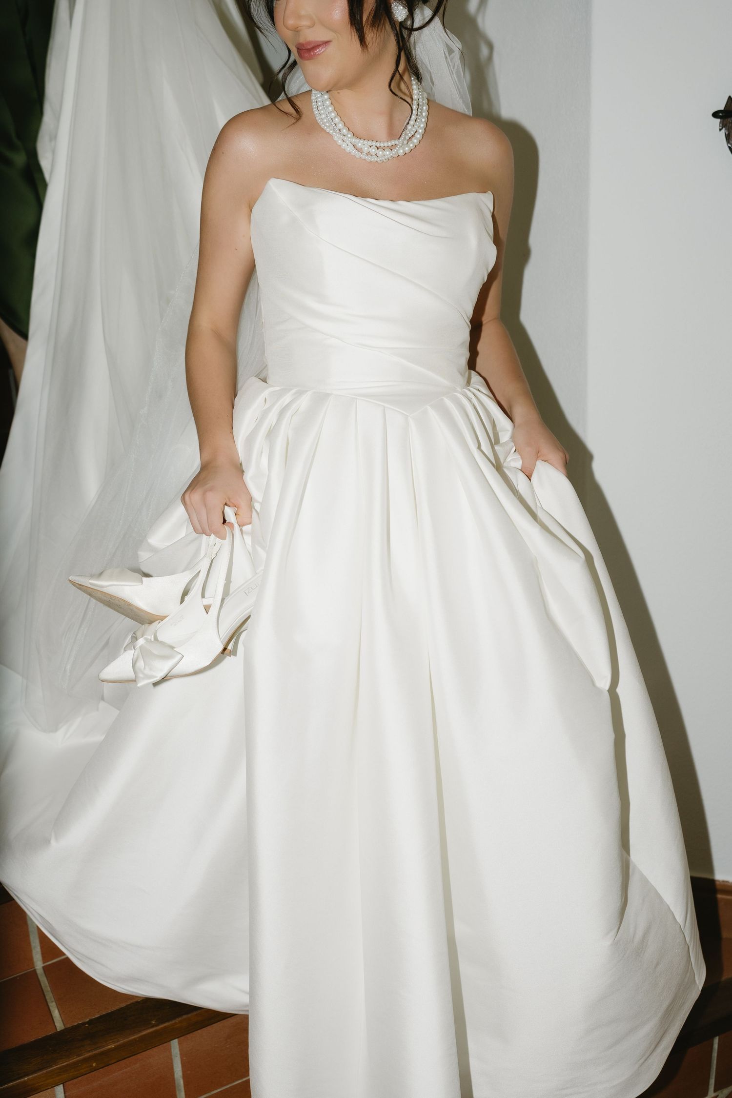 Bride in strapless white ball gown holding heels with pearl necklace and elegant draping.