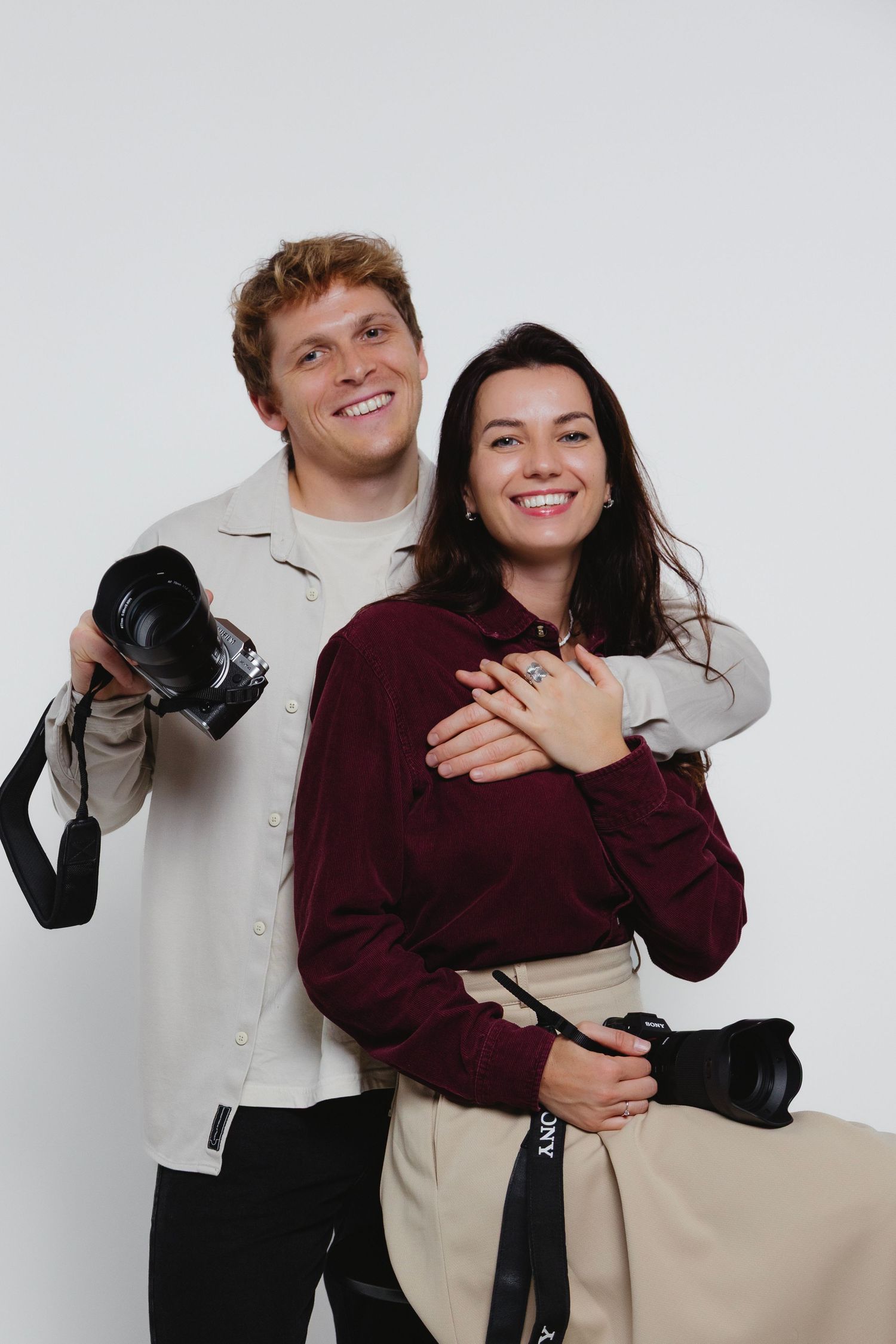 Cheerful couple posing with professional cameras, woman in burgundy embraced from behind by smiling man.