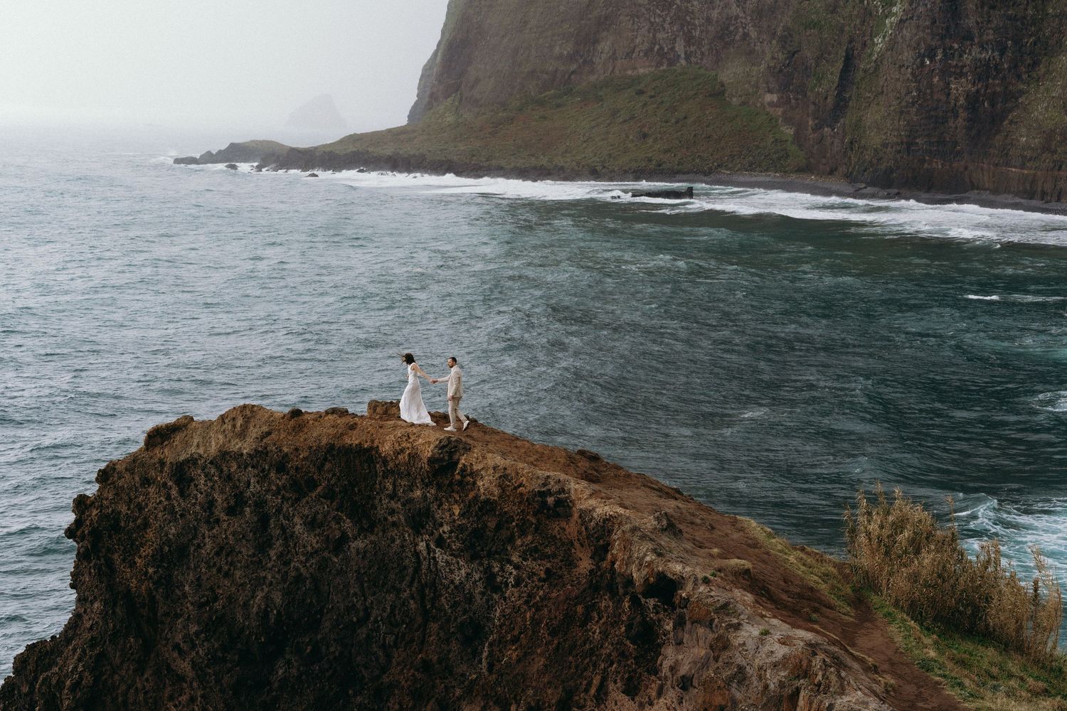 Couple standing on a rugged cliff edge overlooking a dramatic stormy ocean.