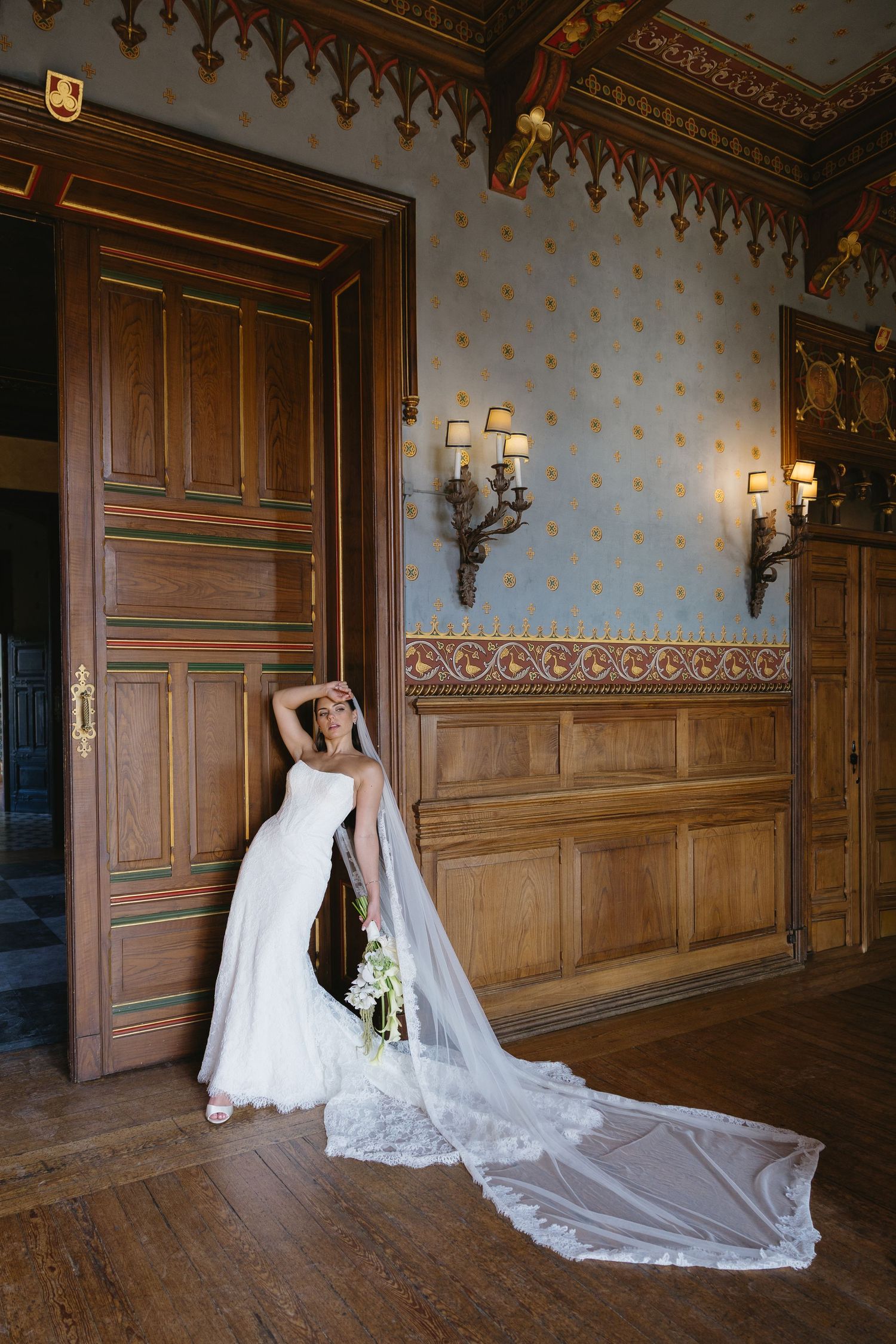 Bride in white gown with long lace veil poses by ornate wooden doors in an elegant historic room.