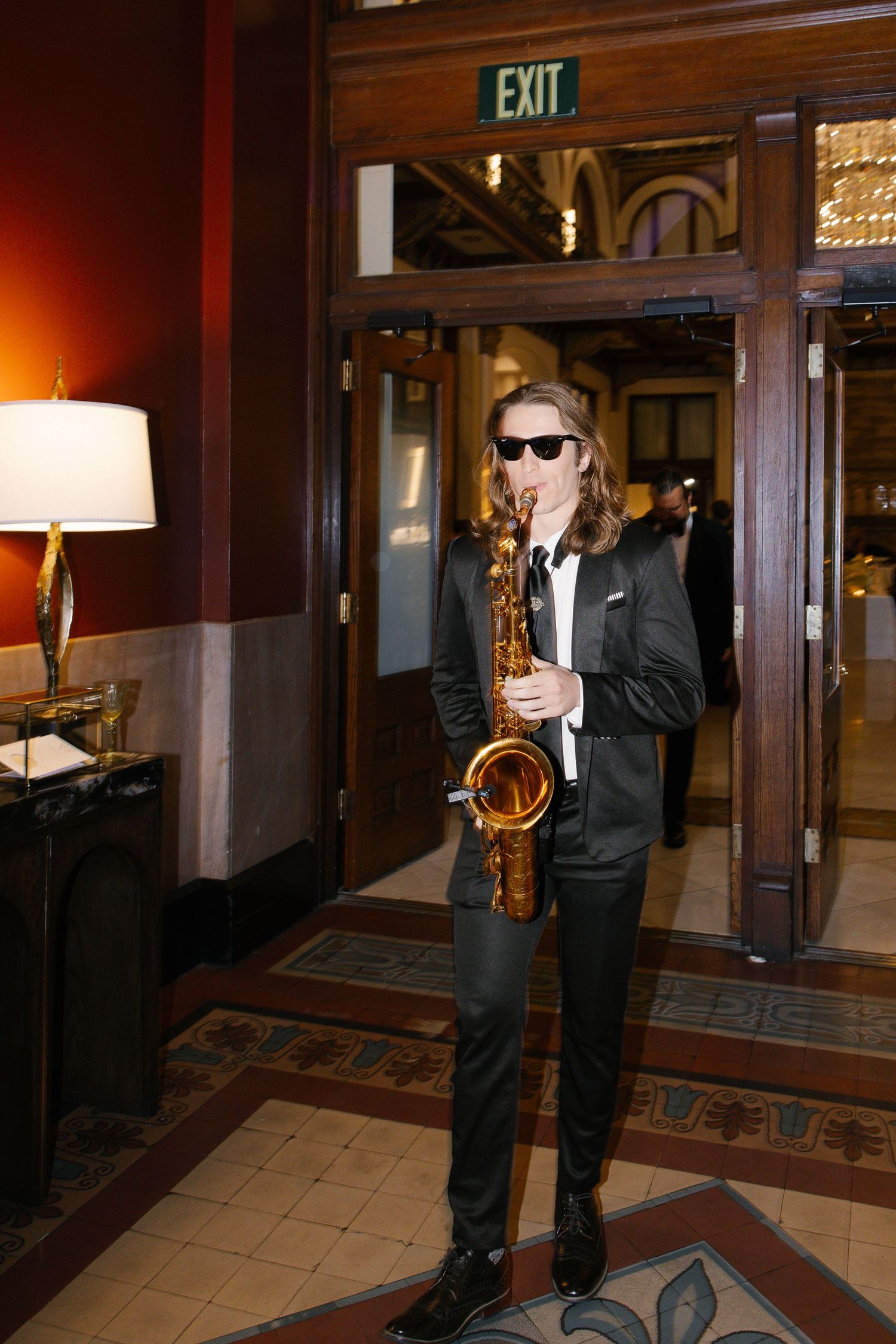 A long-haired musician in a black suit and sunglasses plays saxophone in an elegant hotel lobby near an exit sign.