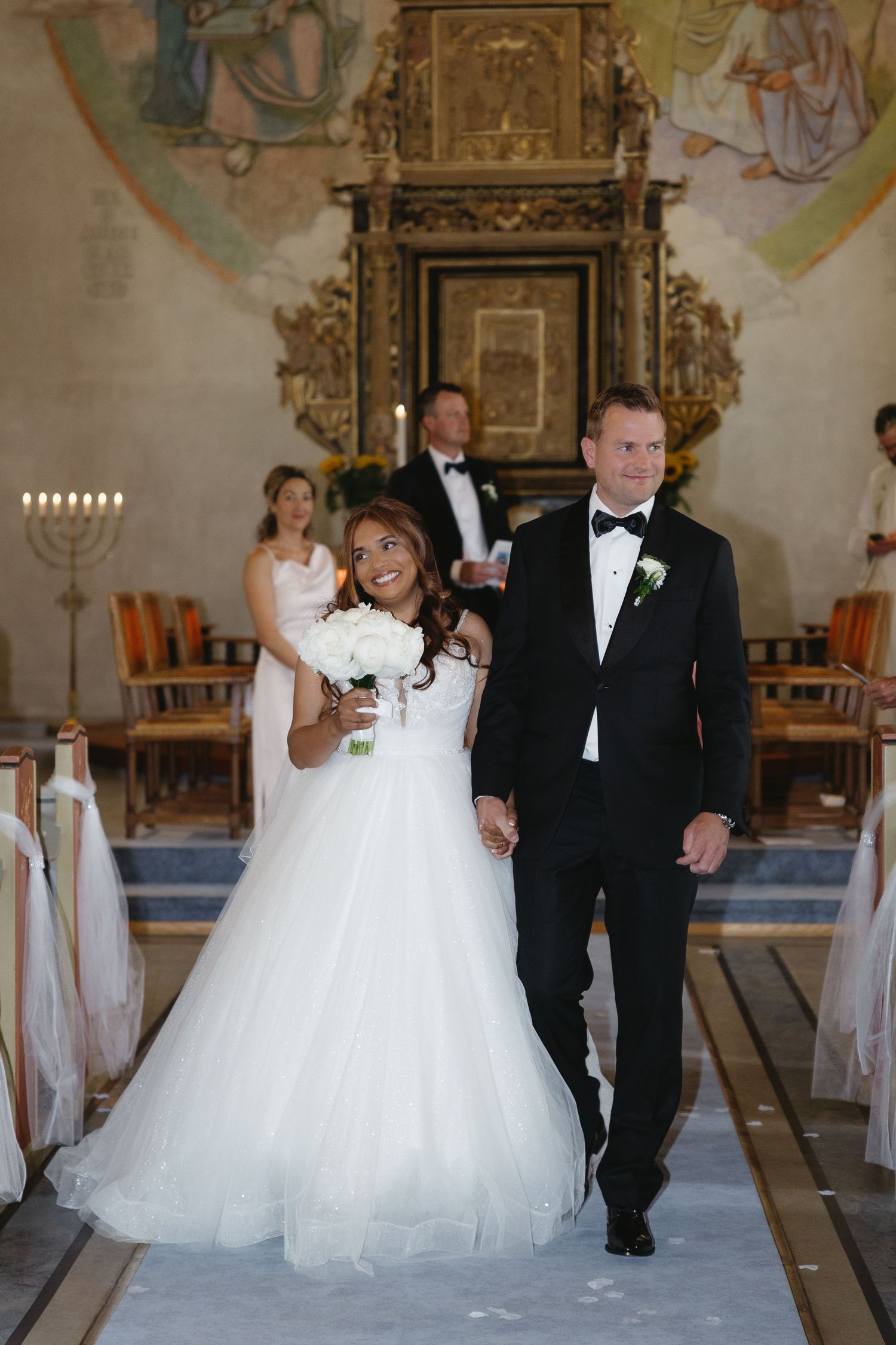 Bride in white ball gown and groom in black tuxedo walk down church aisle after wedding ceremony.