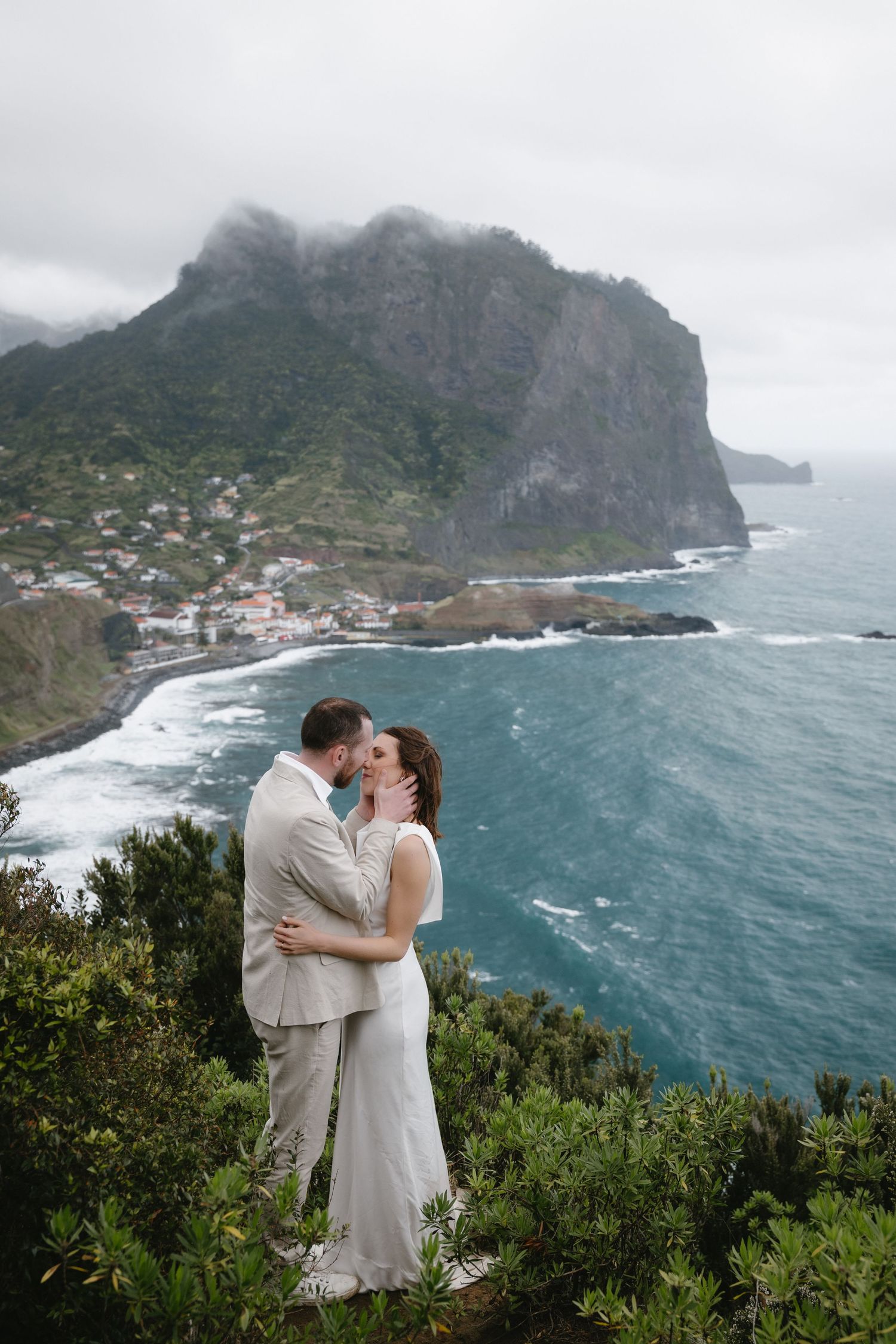 Wedding couple kissing on a lush clifftop with stunning turquoise ocean and misty mountain backdrop in Madeira.