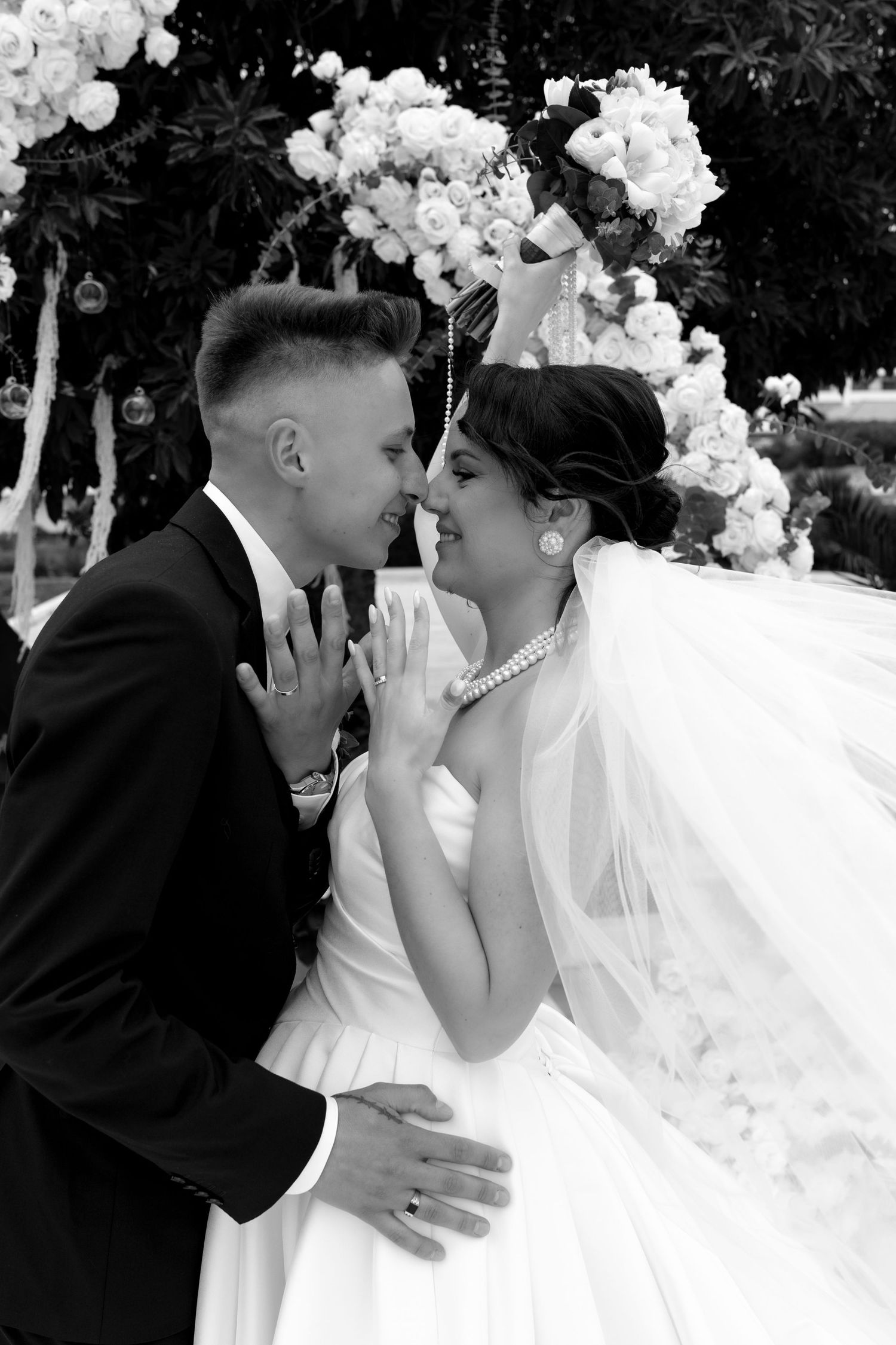 Black and white romantic moment of bride and groom nearly kissing under floral wedding arch.
