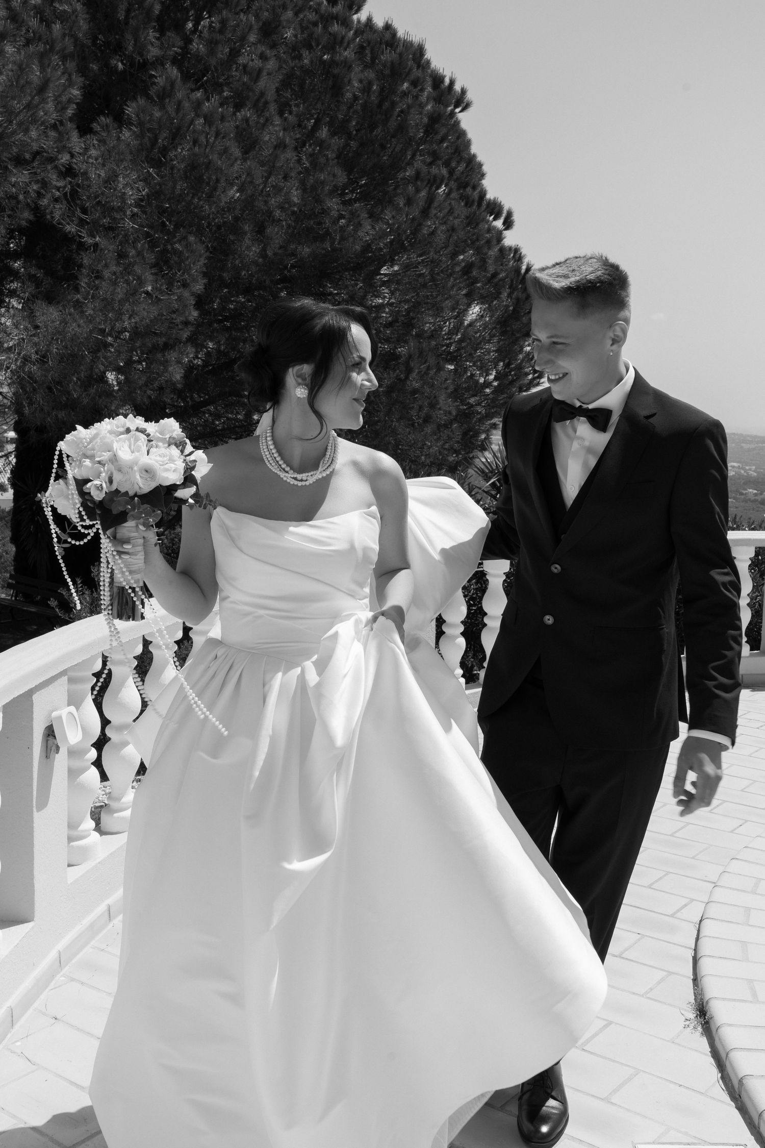 Black and white photo of a smiling bride and groom walking together on a terrace, bride holding a bouquet.