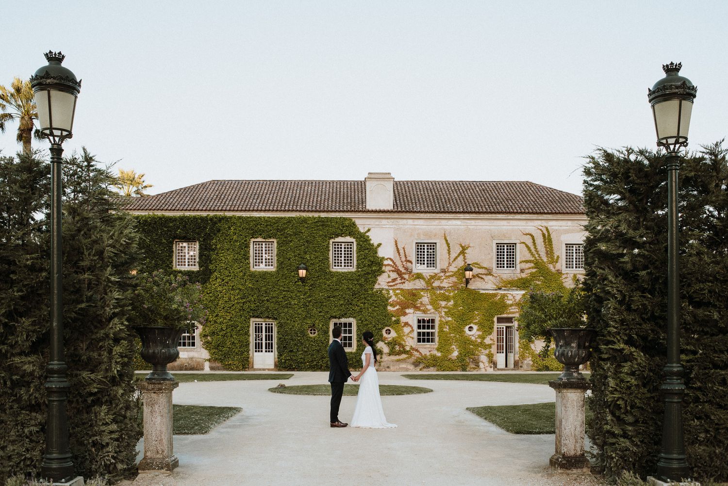 Bride and groom posing in front of an ivy-covered historic estate with elegant lamp posts at dusk.