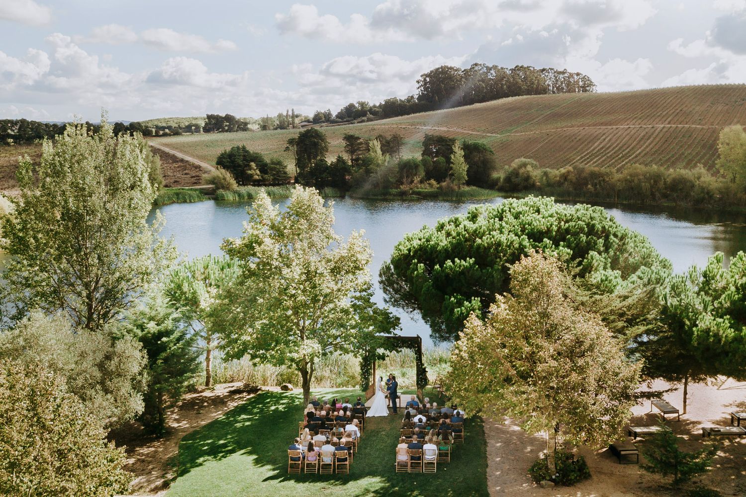 Aerial view of outdoor wedding ceremony by a serene lake surrounded by lush greenery.