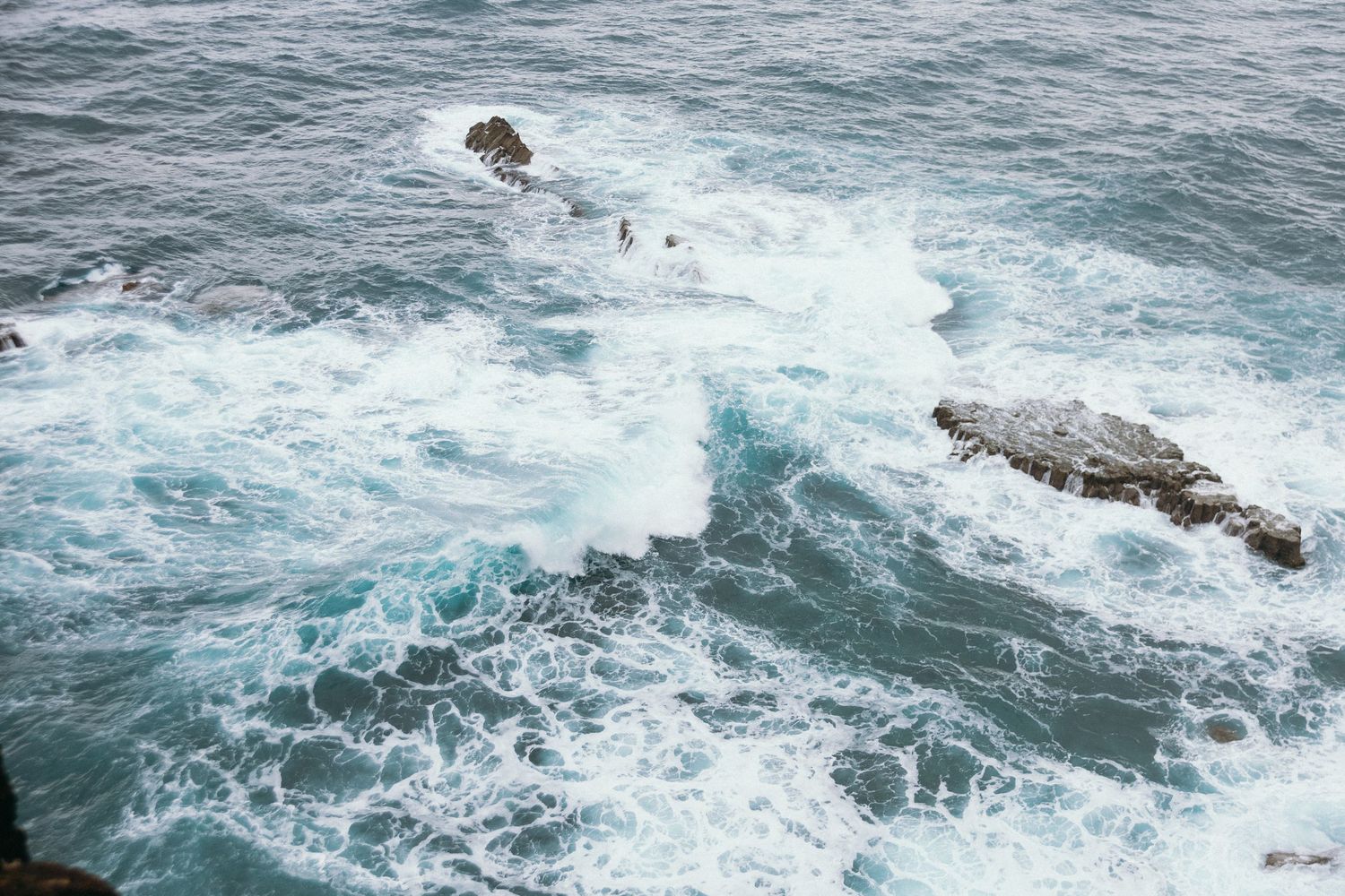 Powerful ocean waves crash against rocky outcroppings creating white sea foam in turquoise blue water.