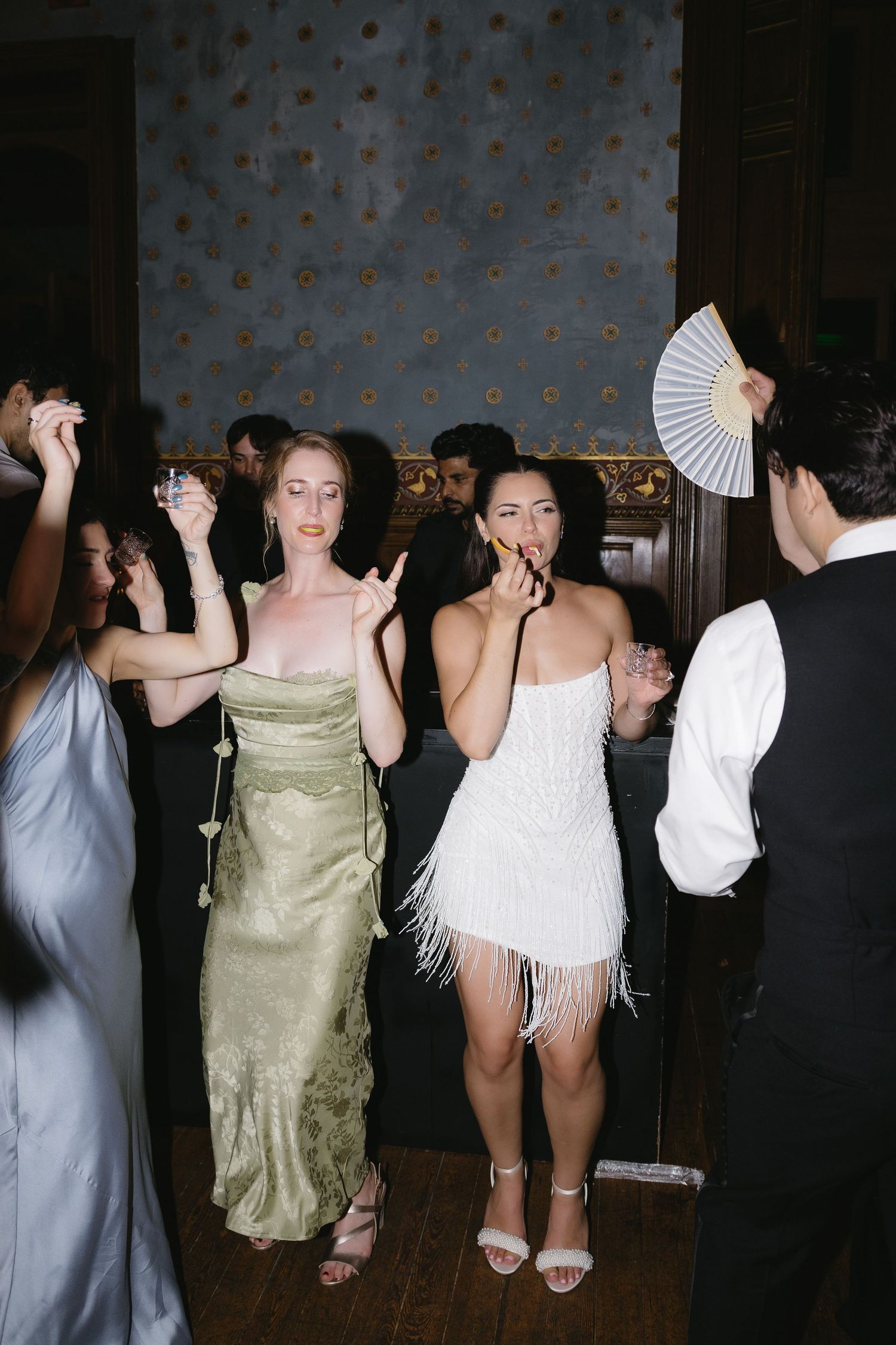 Two women dancing at elegant party, one in green satin gown, other in white fringe mini dress, in ornate ballroom.