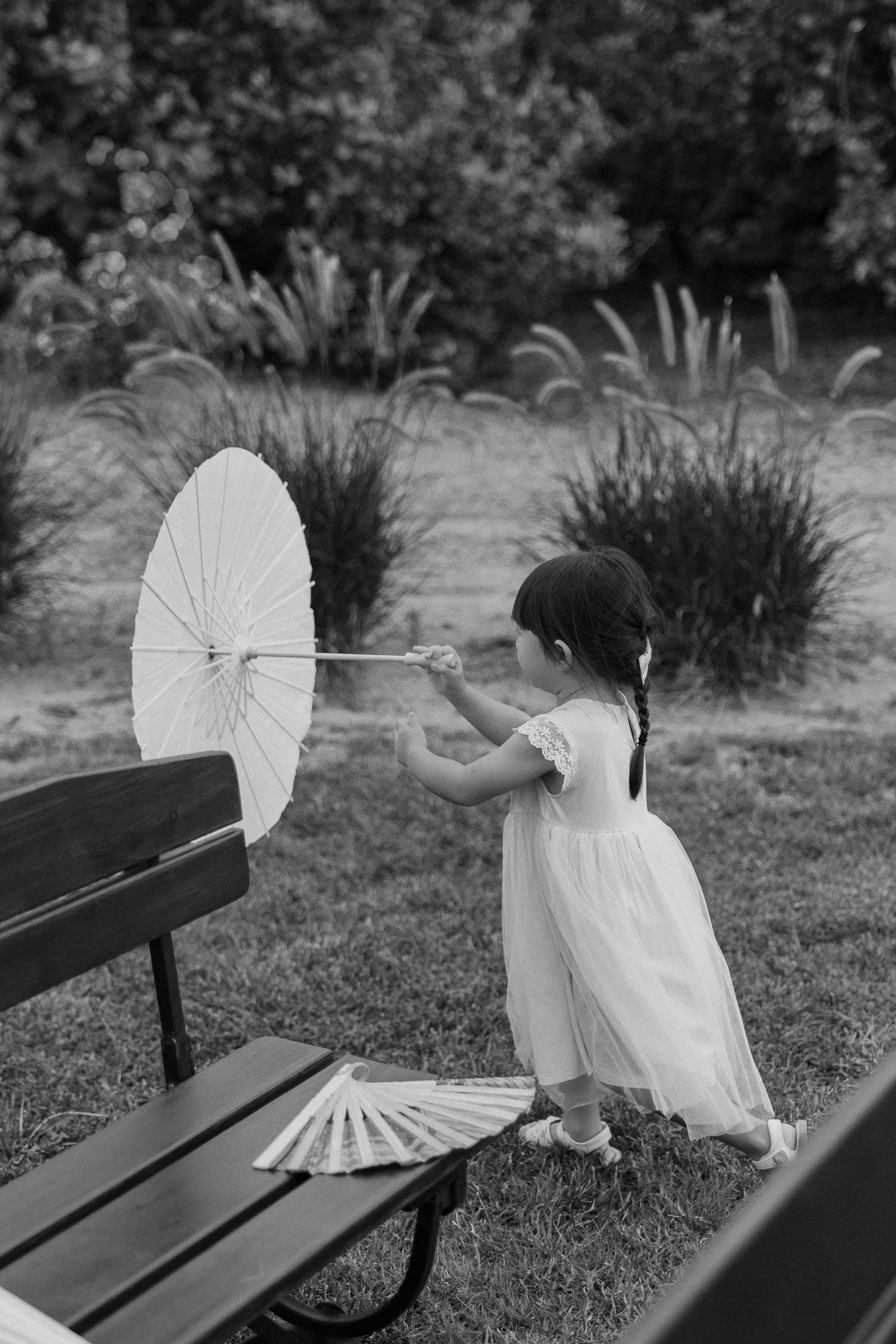 Black and white photo of a young flower girl in white dress holding a parasol beside a wooden bench in a garden.