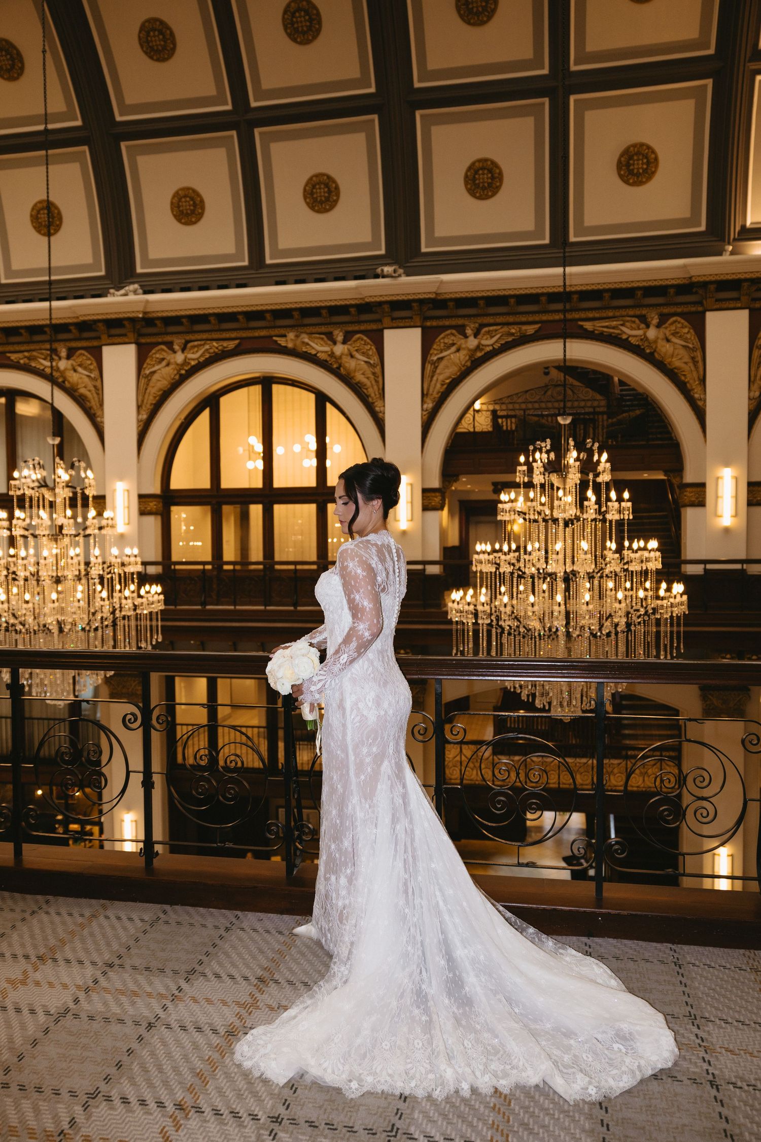 Bride in elegant lace wedding gown with train poses in ornate ballroom with crystal chandeliers.
