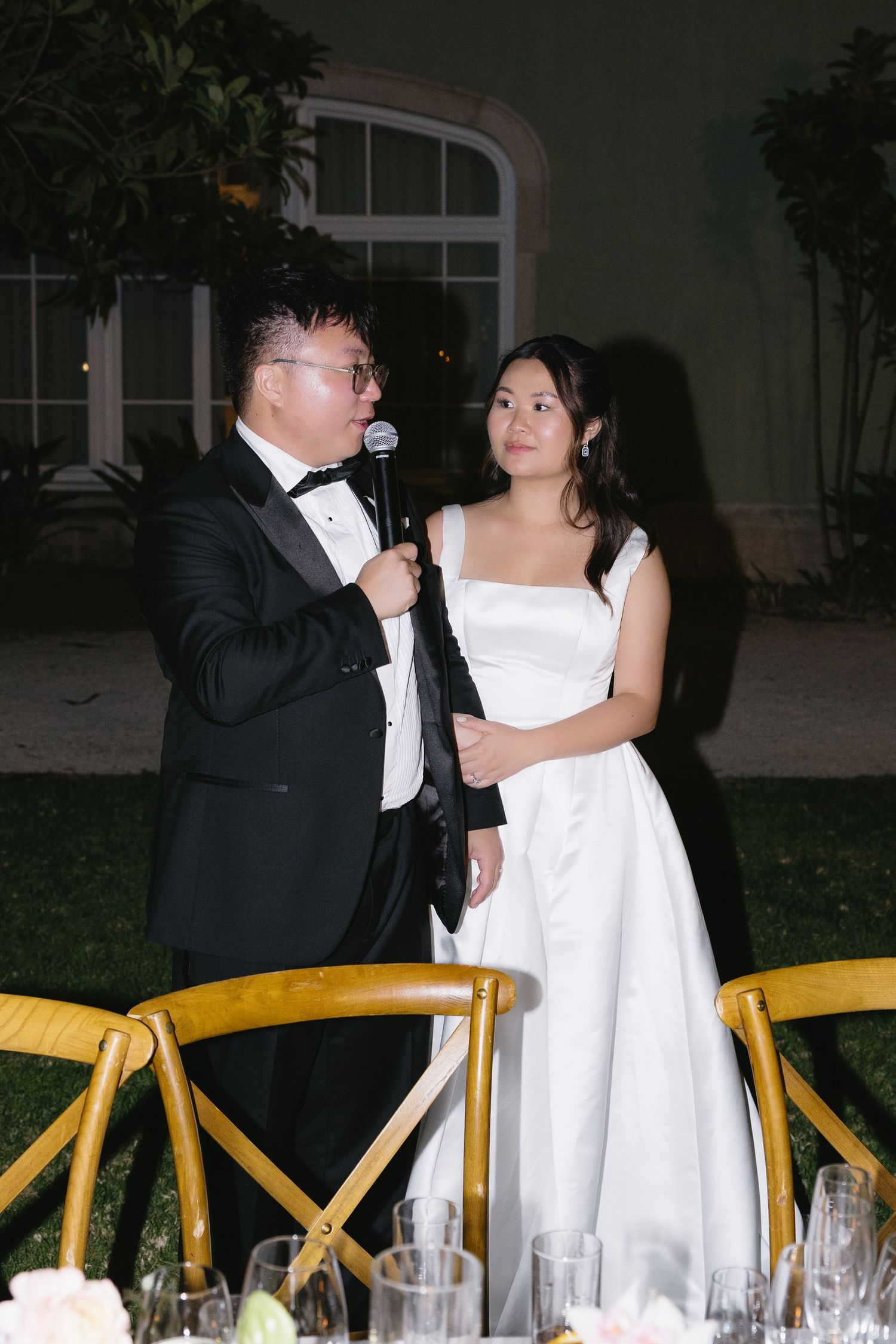 Bride in white gown and groom in tuxedo sharing a microphone during wedding reception speech at elegant outdoor venue.