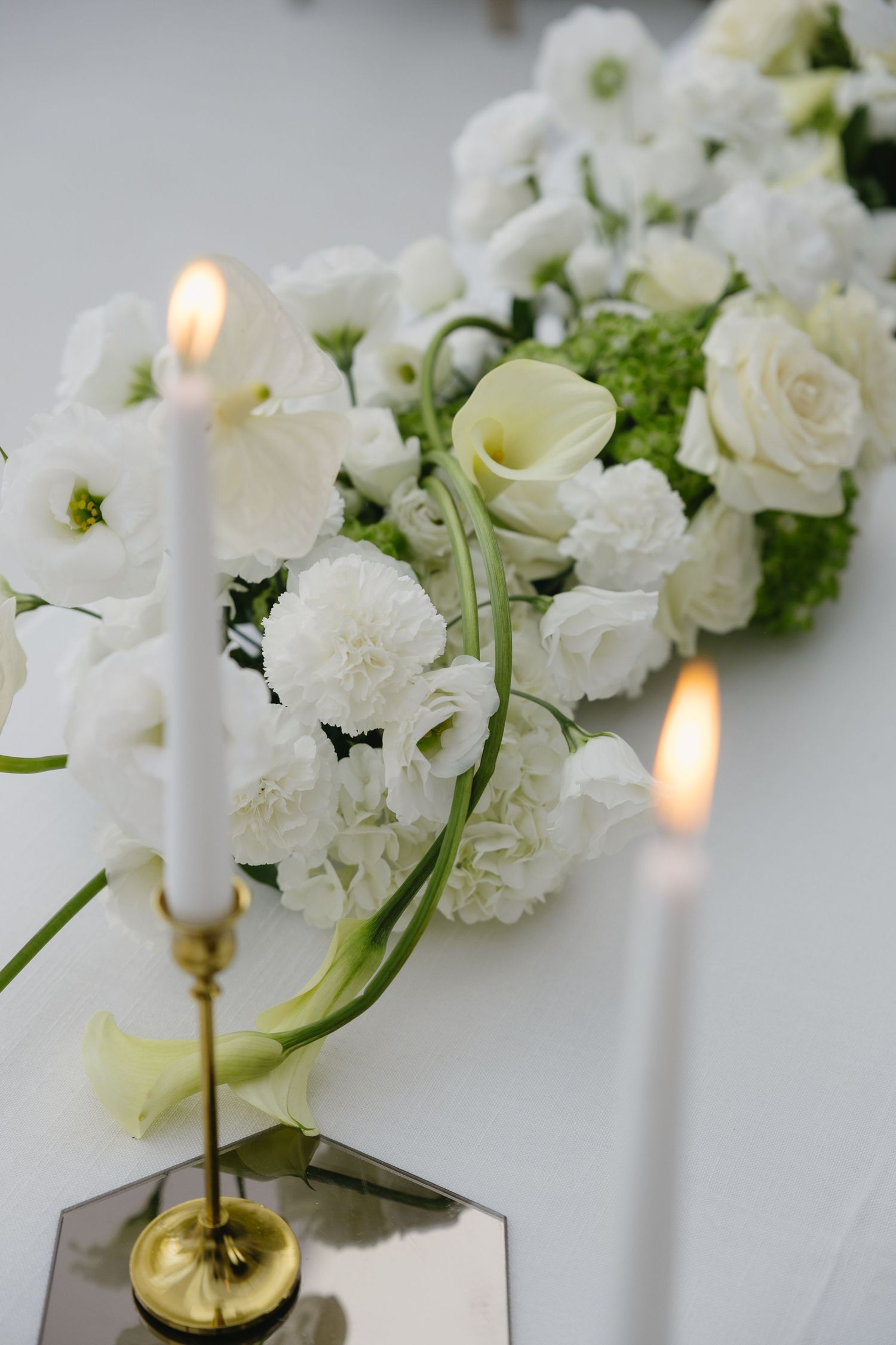 Elegant wedding table with white floral arrangement of roses and carnations flanked by lit candles in gold holders.