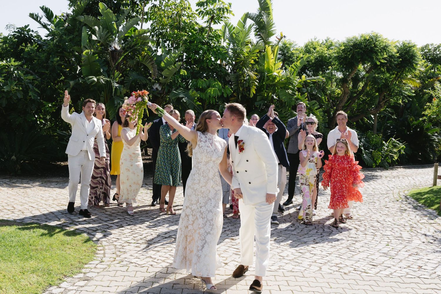 Newlyweds celebrate with guests on cobblestone path, bride raising bouquet joyfully in lush garden setting.
