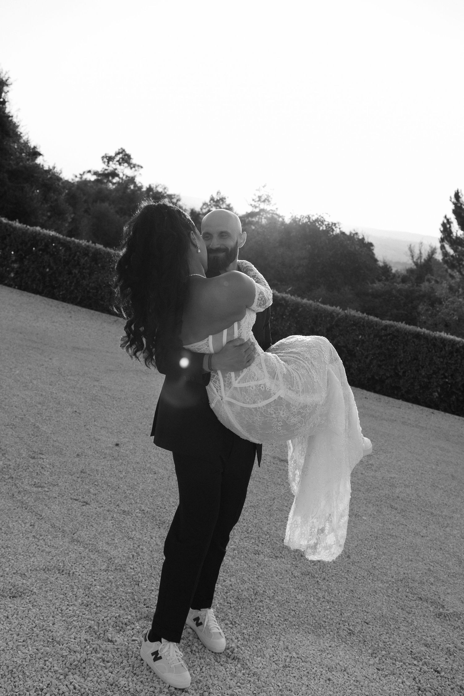 Black and white photo of groom lifting bride in lace wedding dress outdoors at golden hour, romantic moment.